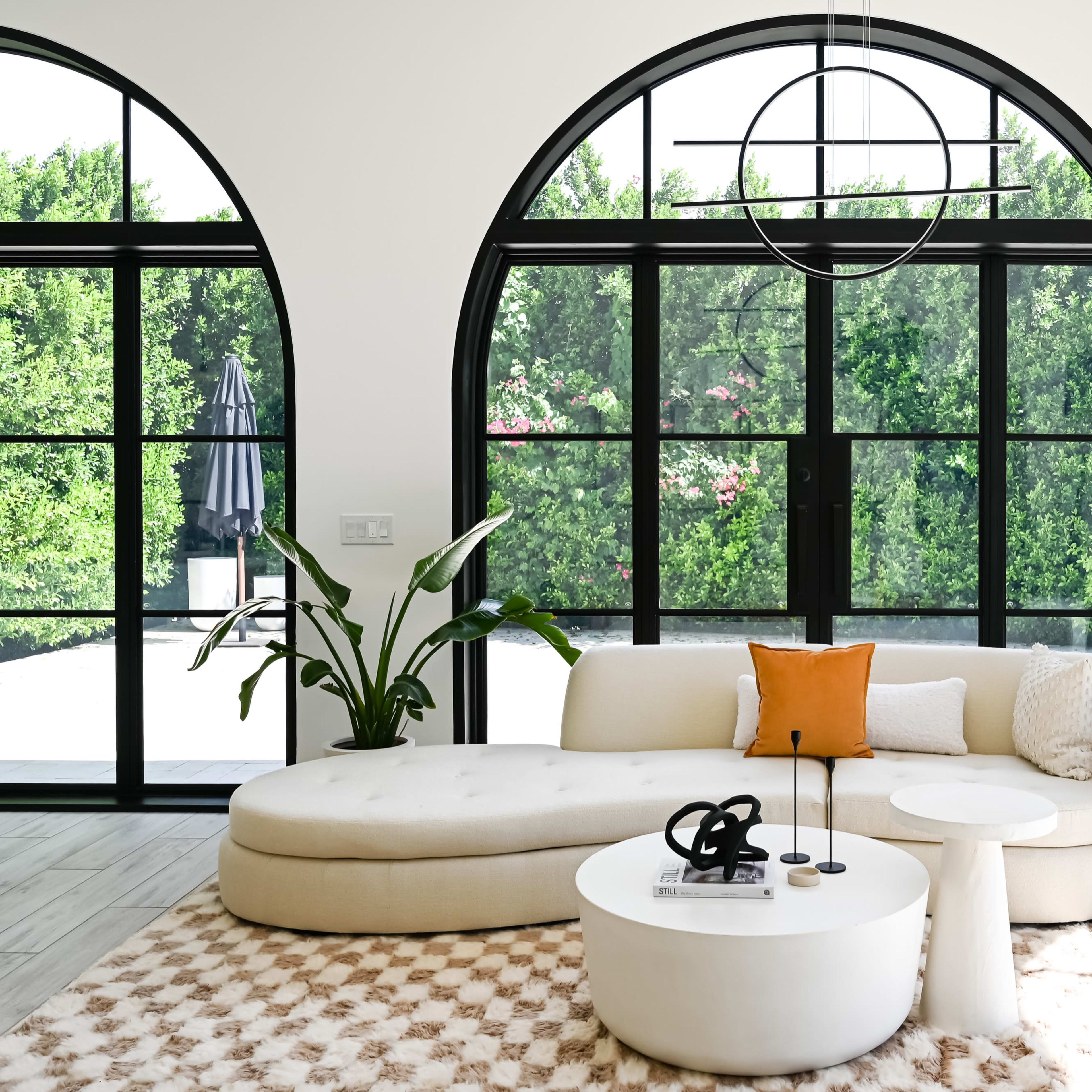 A modern living room featuring a curved sofa, a circular coffee table, and large arched windows that provide views of greenery outside.