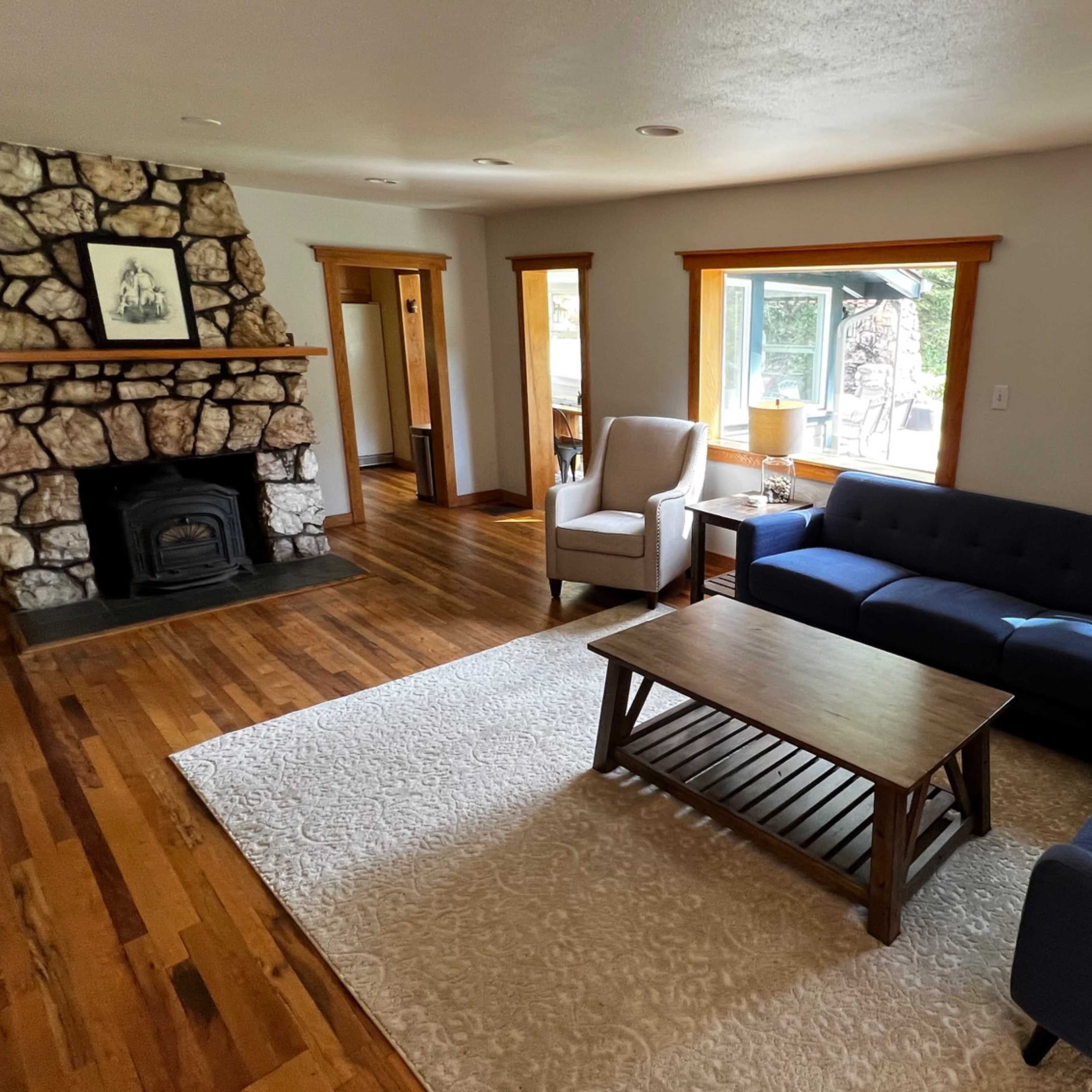 A living room with a stone fireplace, a blue sofa, a wooden coffee table, and large windows allowing natural light to enter.