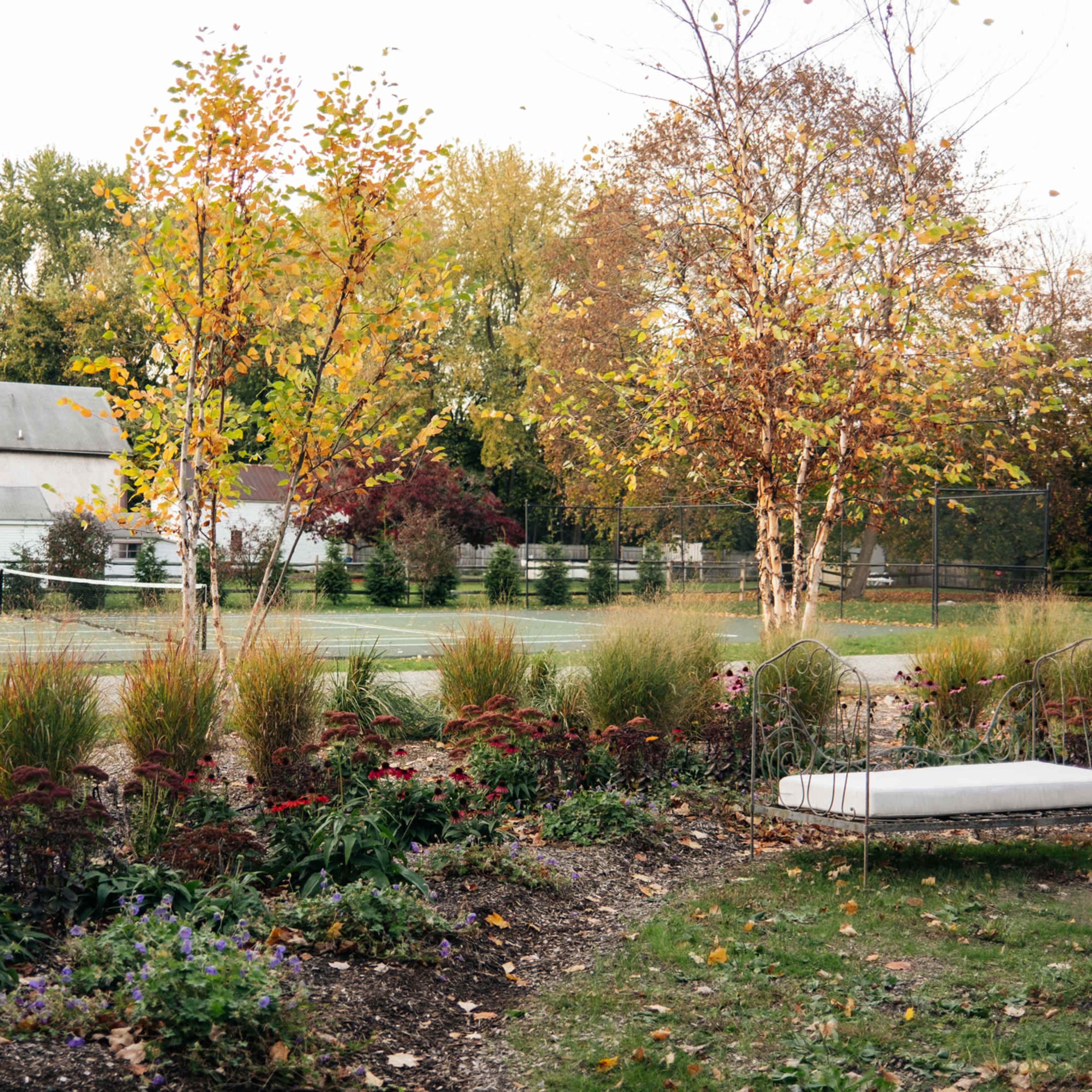 A serene outdoor setting featuring a white swing in a garden bordered by colorful flowers, with a tennis court and a barn in the background.