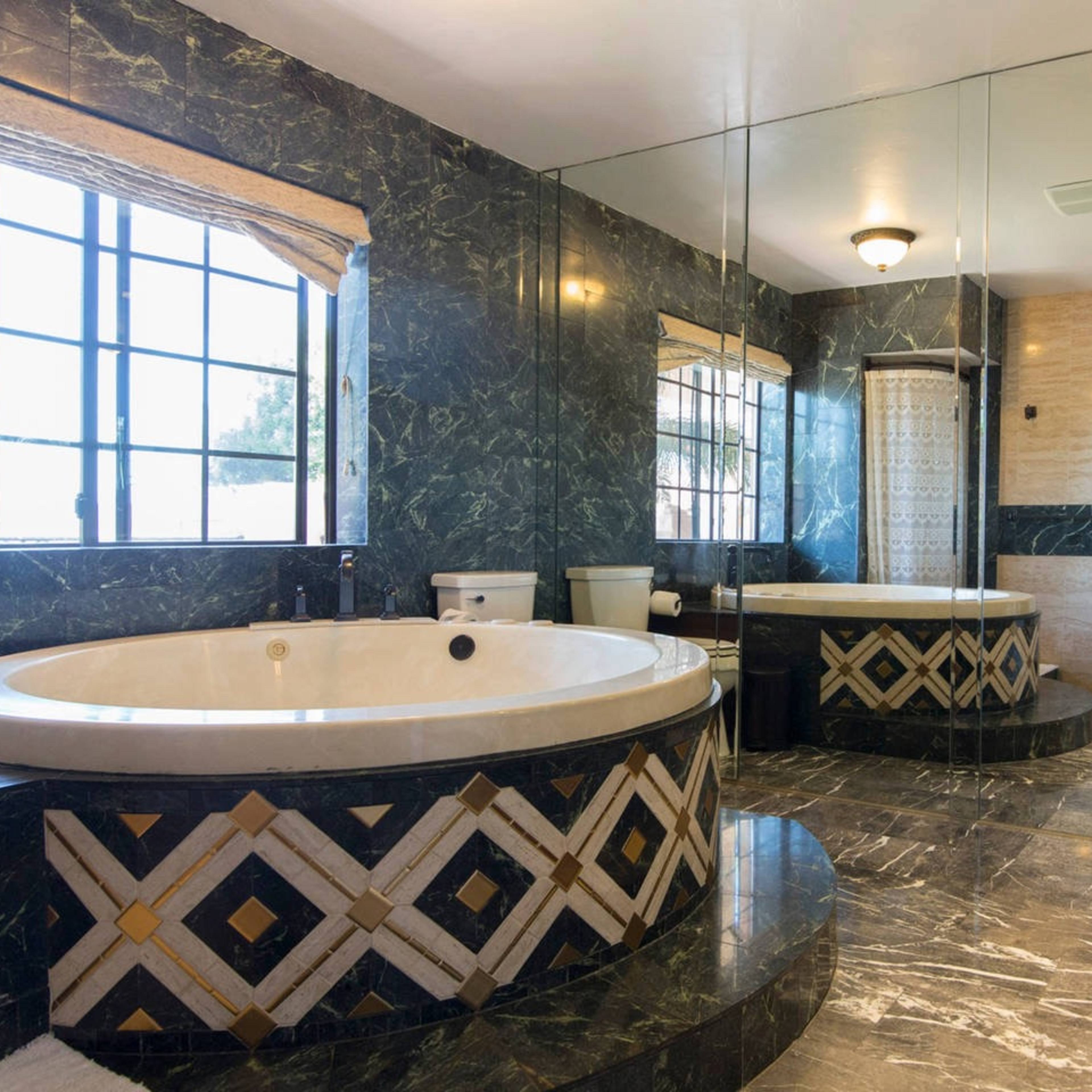 The bathroom features a large circular jacuzzi tub surrounded by marble walls, a separate shower area, and dual sinks with a mirror above each.