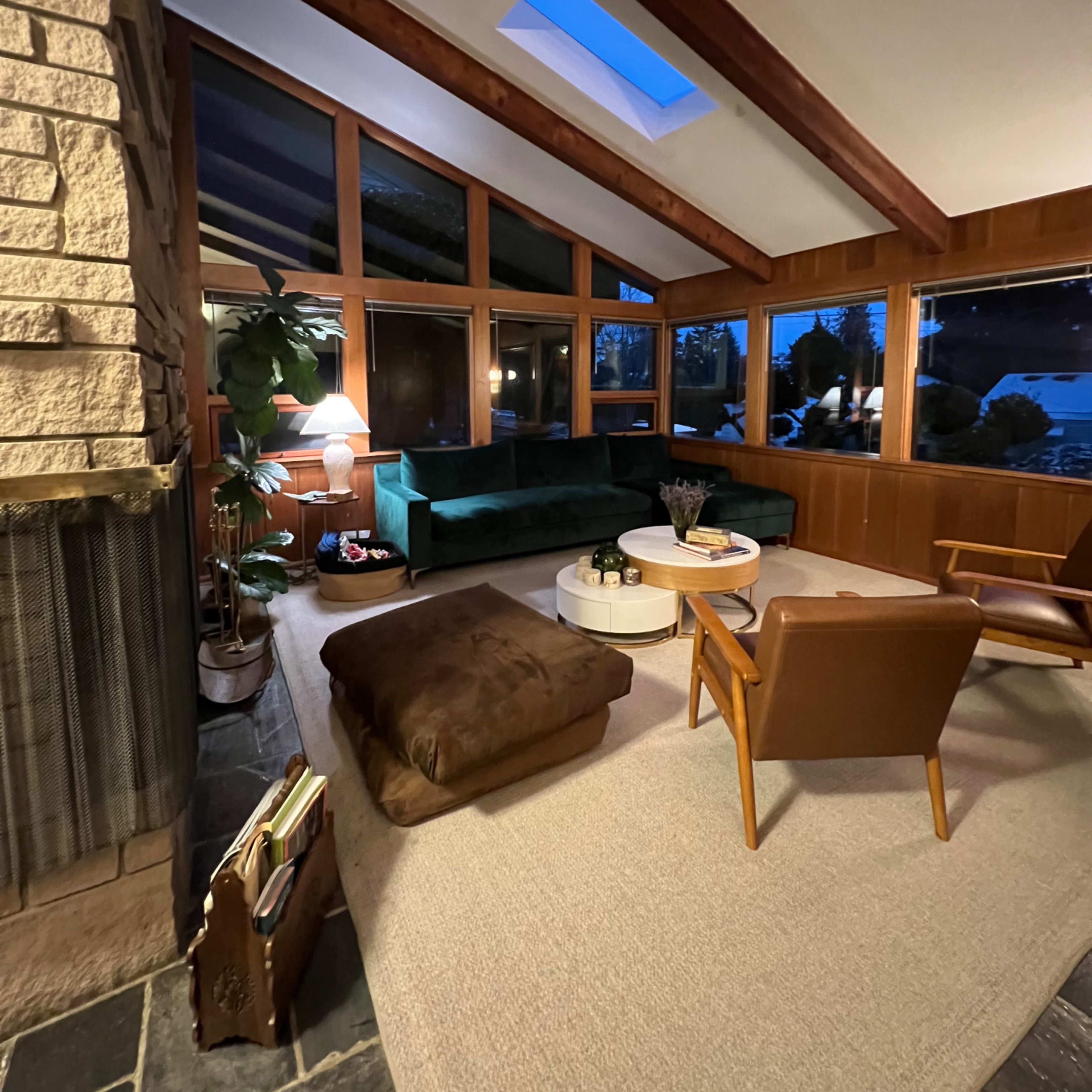 A cozy living room with a stone fireplace, large windows, a green couch, a round coffee table, and a chair, all set on a neutral carpet.