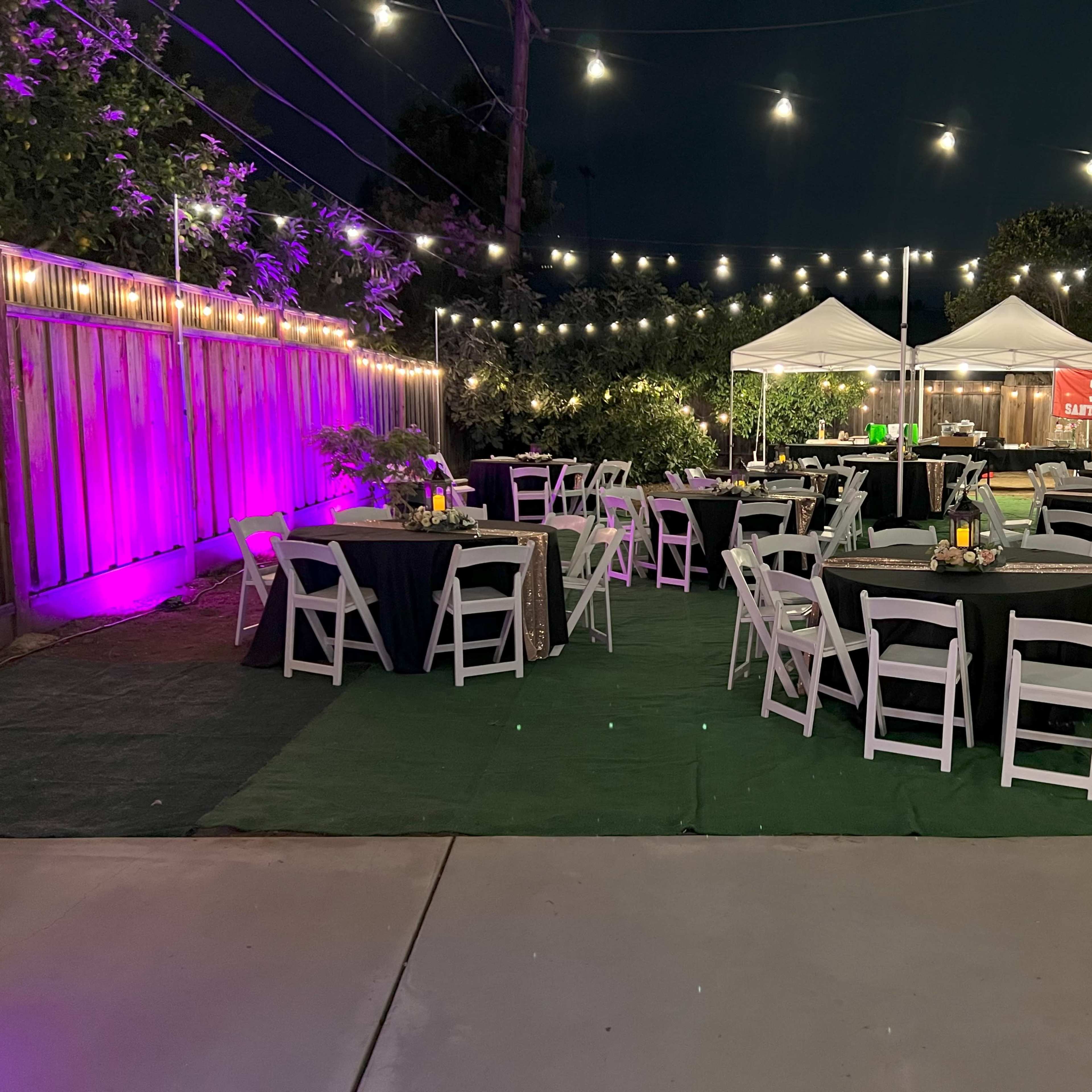 The scene shows a landscaped outdoor event space with tables set for dining, string lights overhead, and canopies in the background.