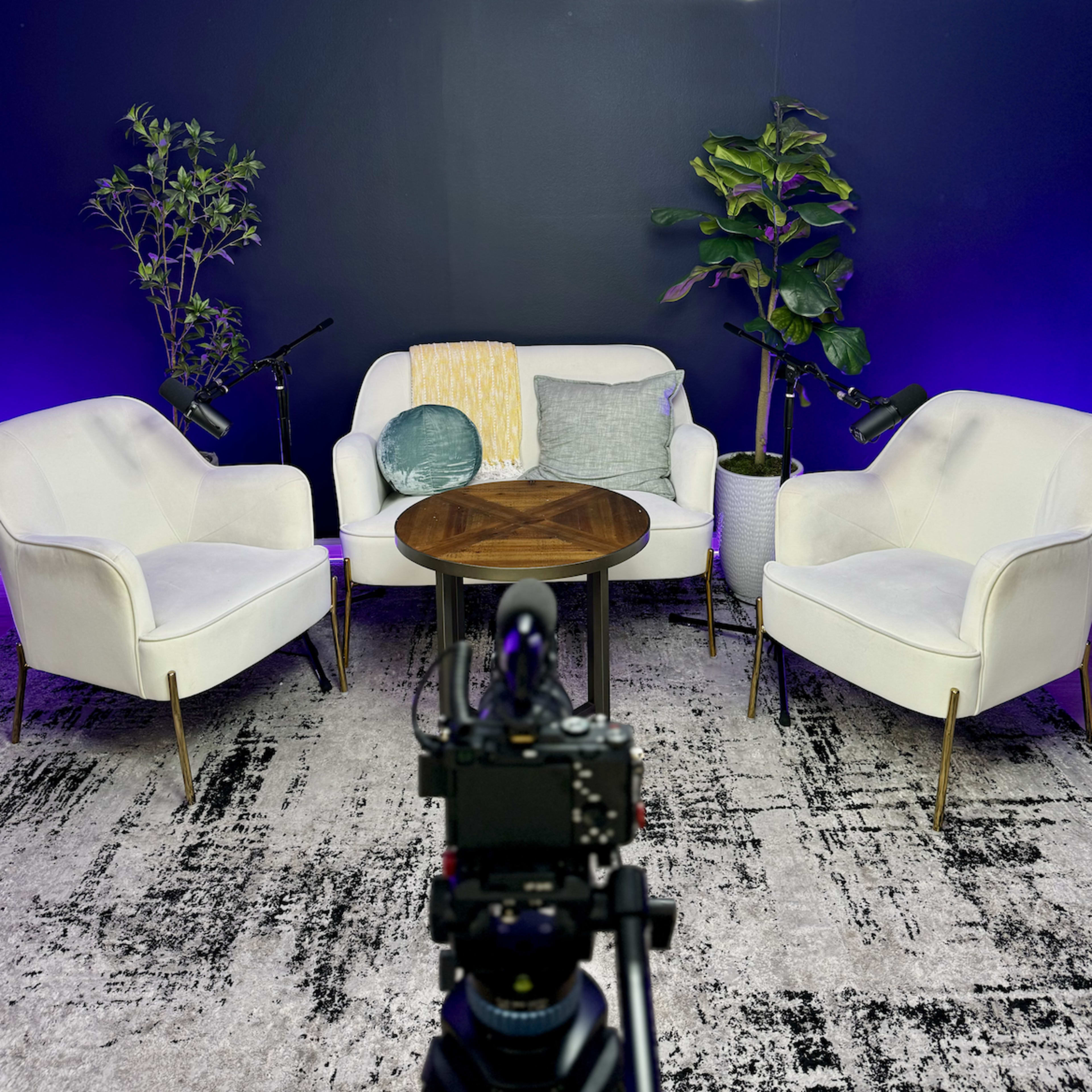 A modern seating area with two white chairs and a round wooden table, surrounded by potted plants against a dark wall.
