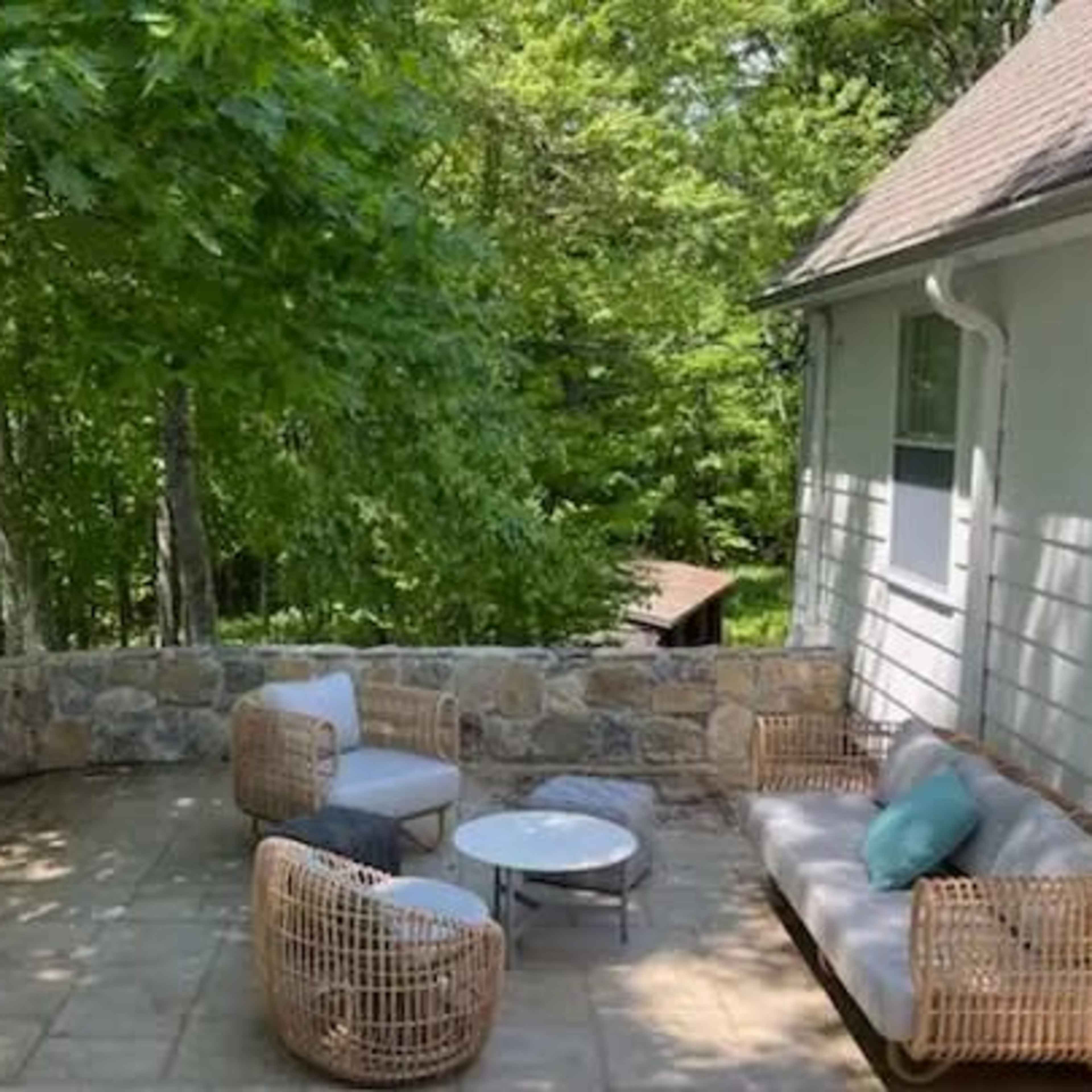 A stone patio with wicker seating and a small table is surrounded by greenery.