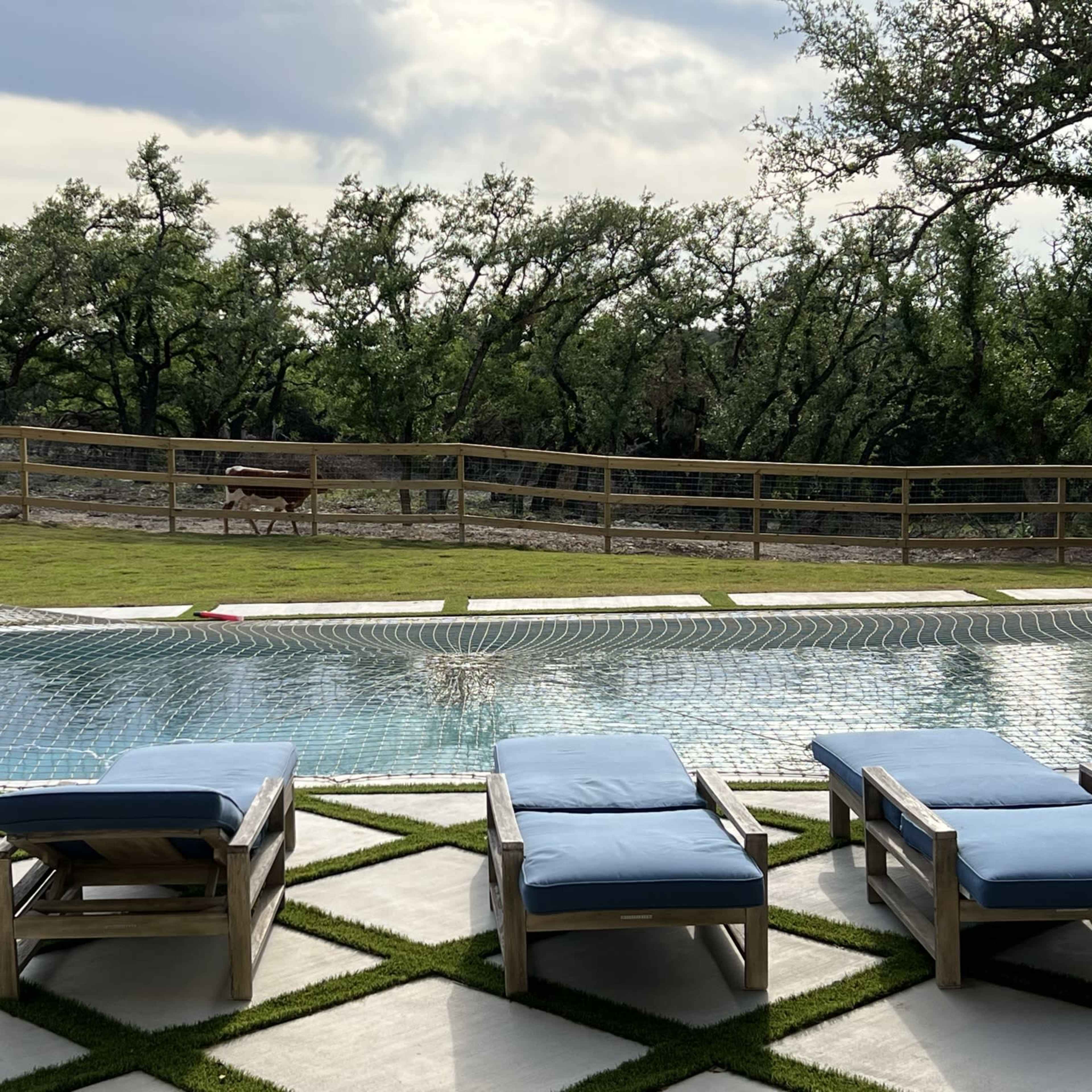 A swimming pool surrounded by lounge chairs, with a grassy area and trees in the background.