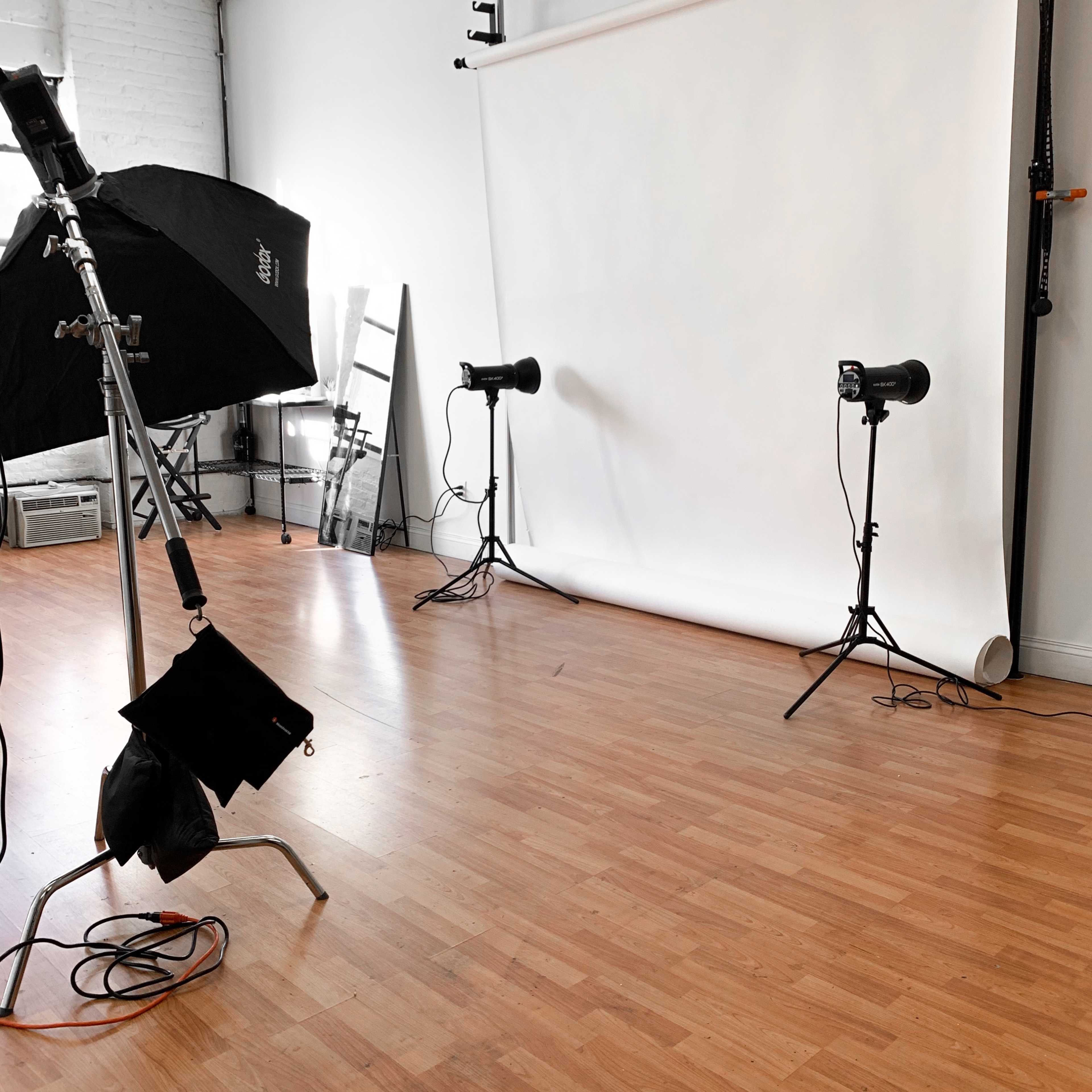 A photography studio with a white backdrop, two studio lights on stands, and a softbox set up on the floor.