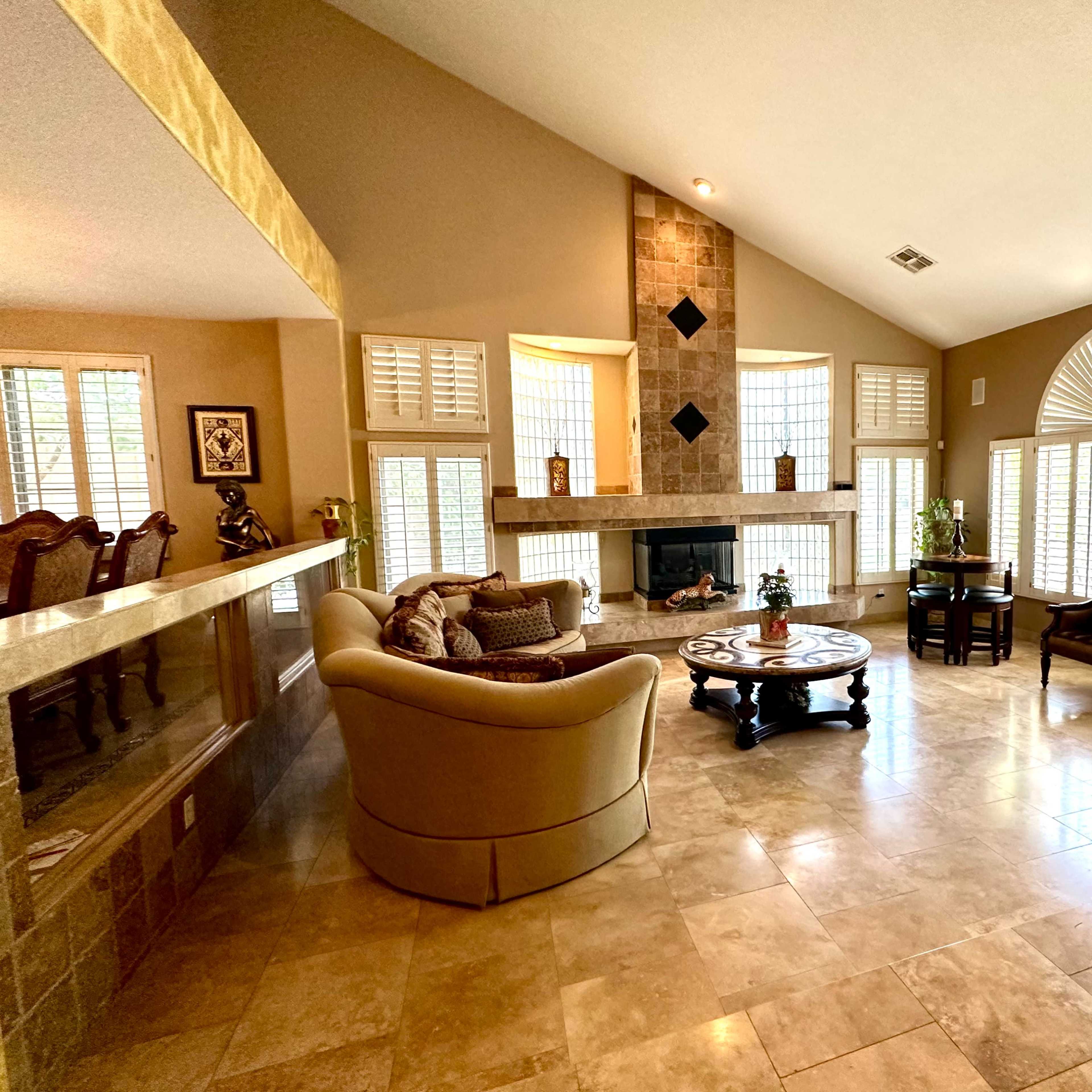 A spacious living room features a central fireplace, large windows with shutters, and a mix of comfortable seating around a round coffee table.