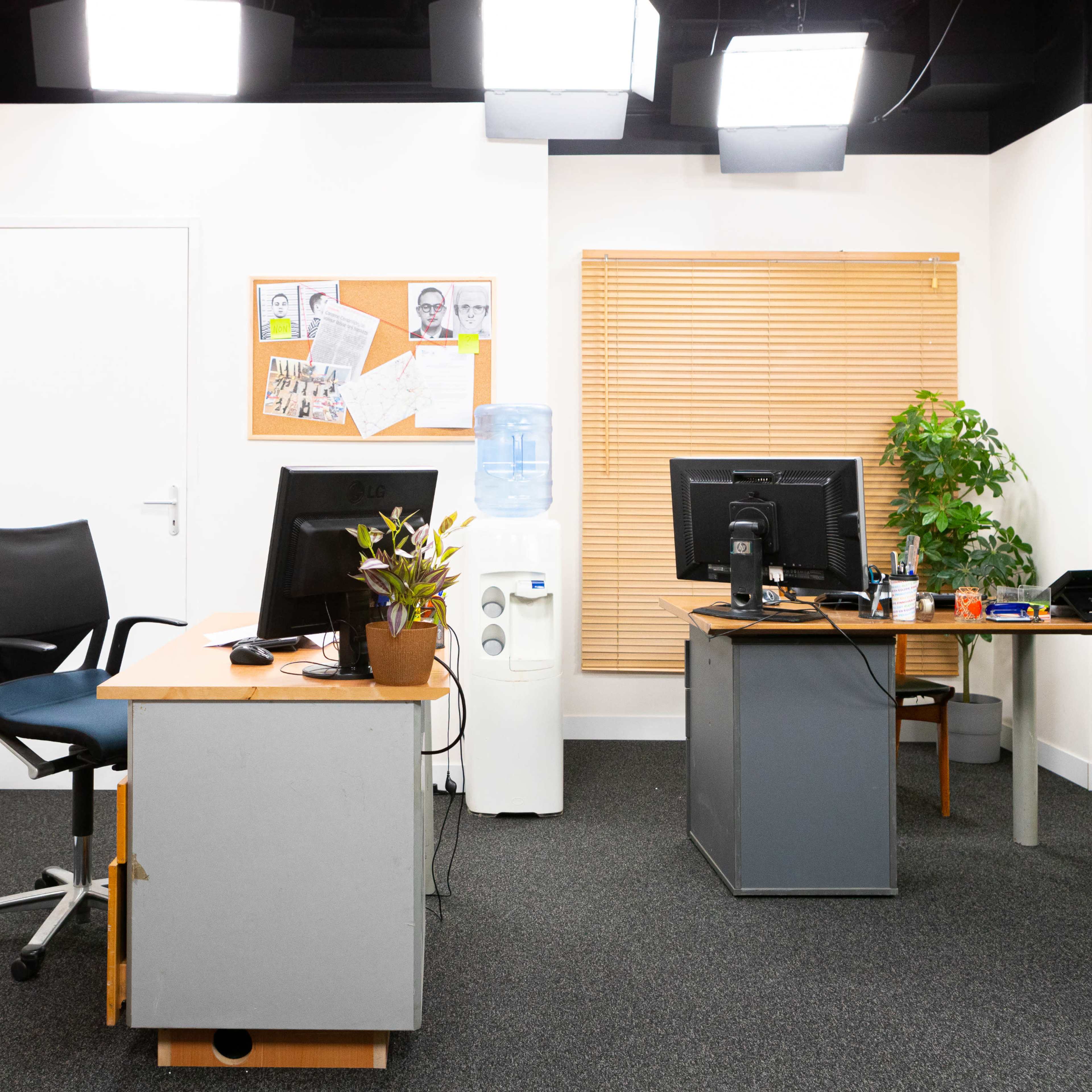 A simple office setup with two desks, a water cooler, a plant, and a schedule board mounted on the wall.