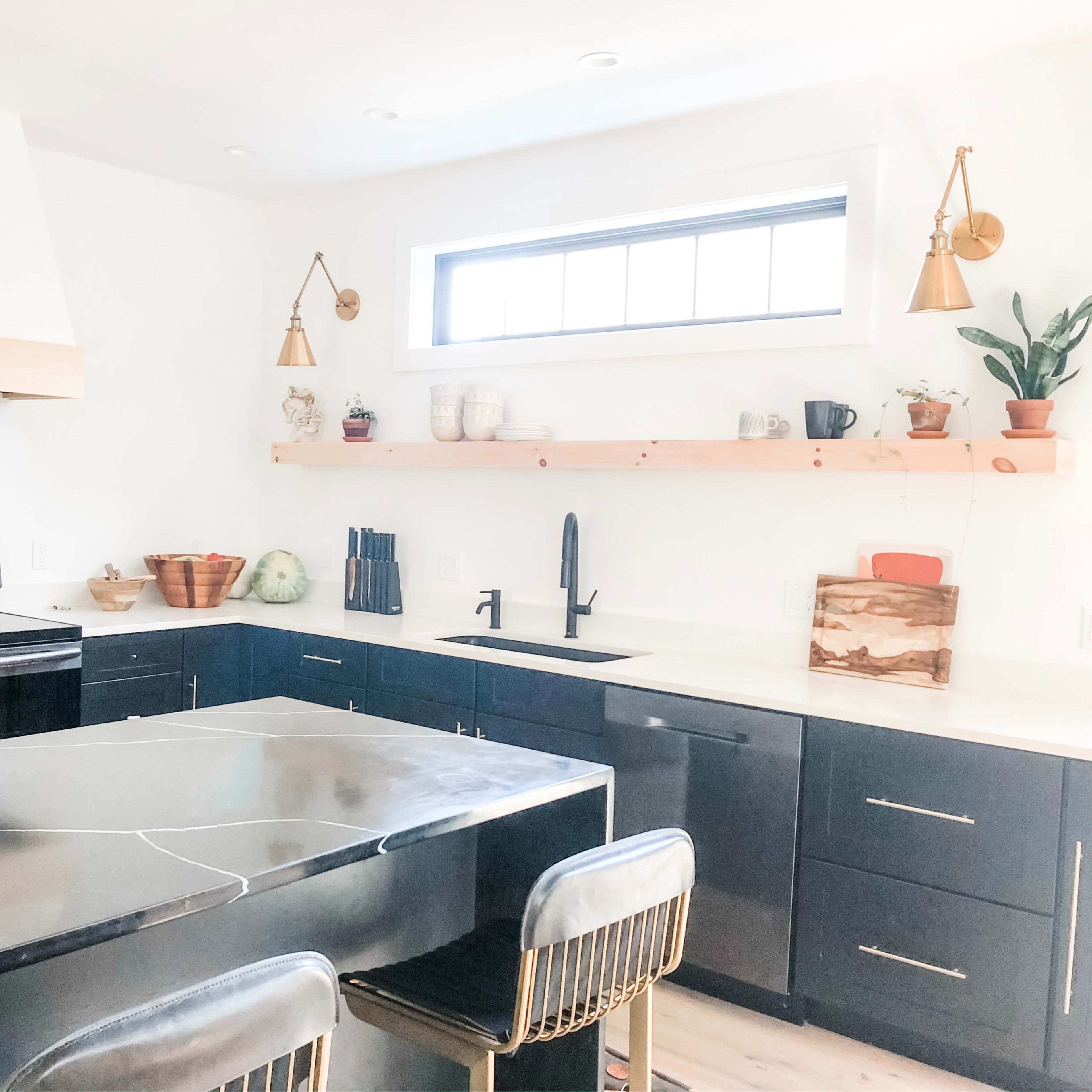 A modern kitchen features black cabinets, a central island with a black marble countertop, and wooden shelving above a stainless steel sink.