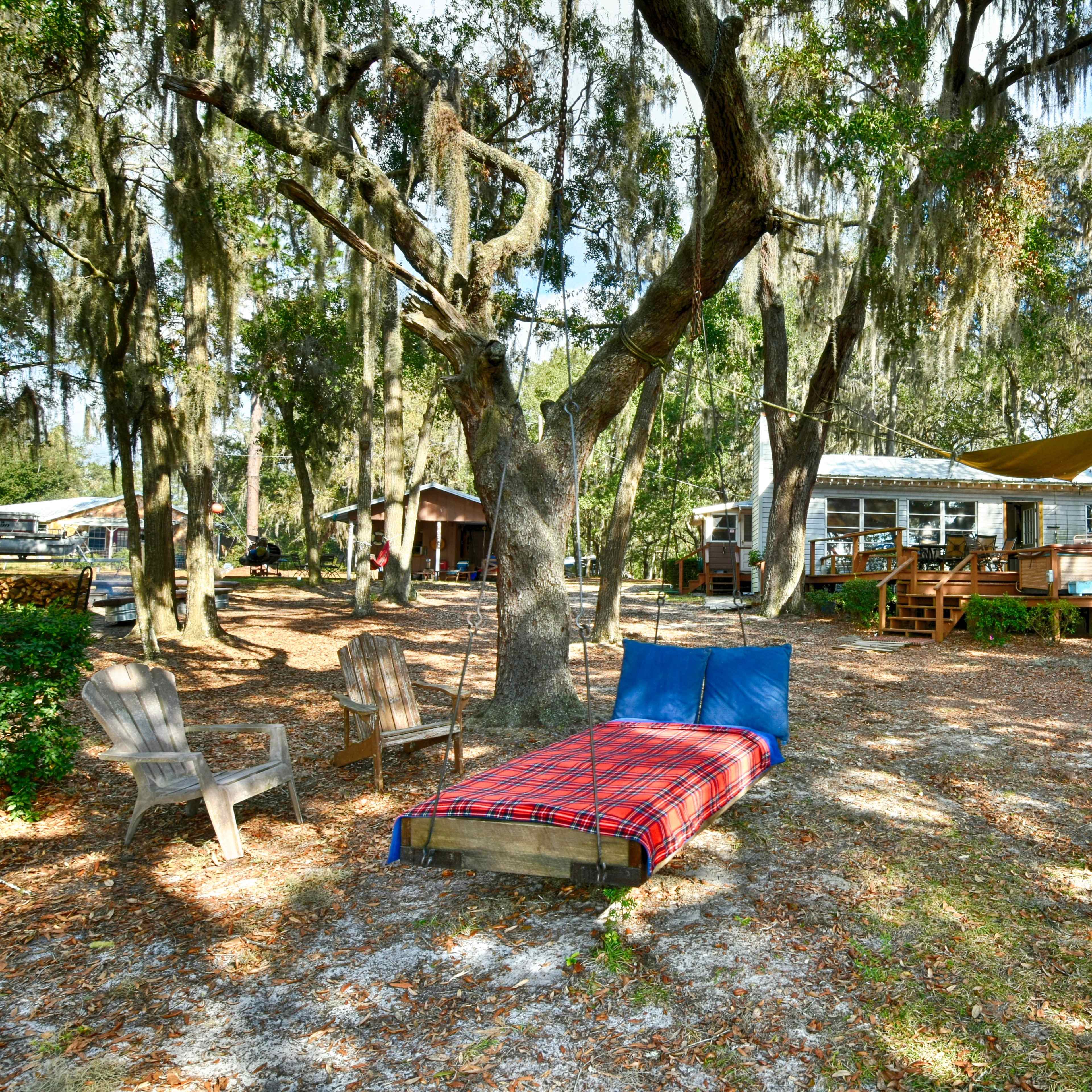 A wooden bed with a blue mattress is positioned in a grassy area near a house surrounded by trees with Spanish moss.