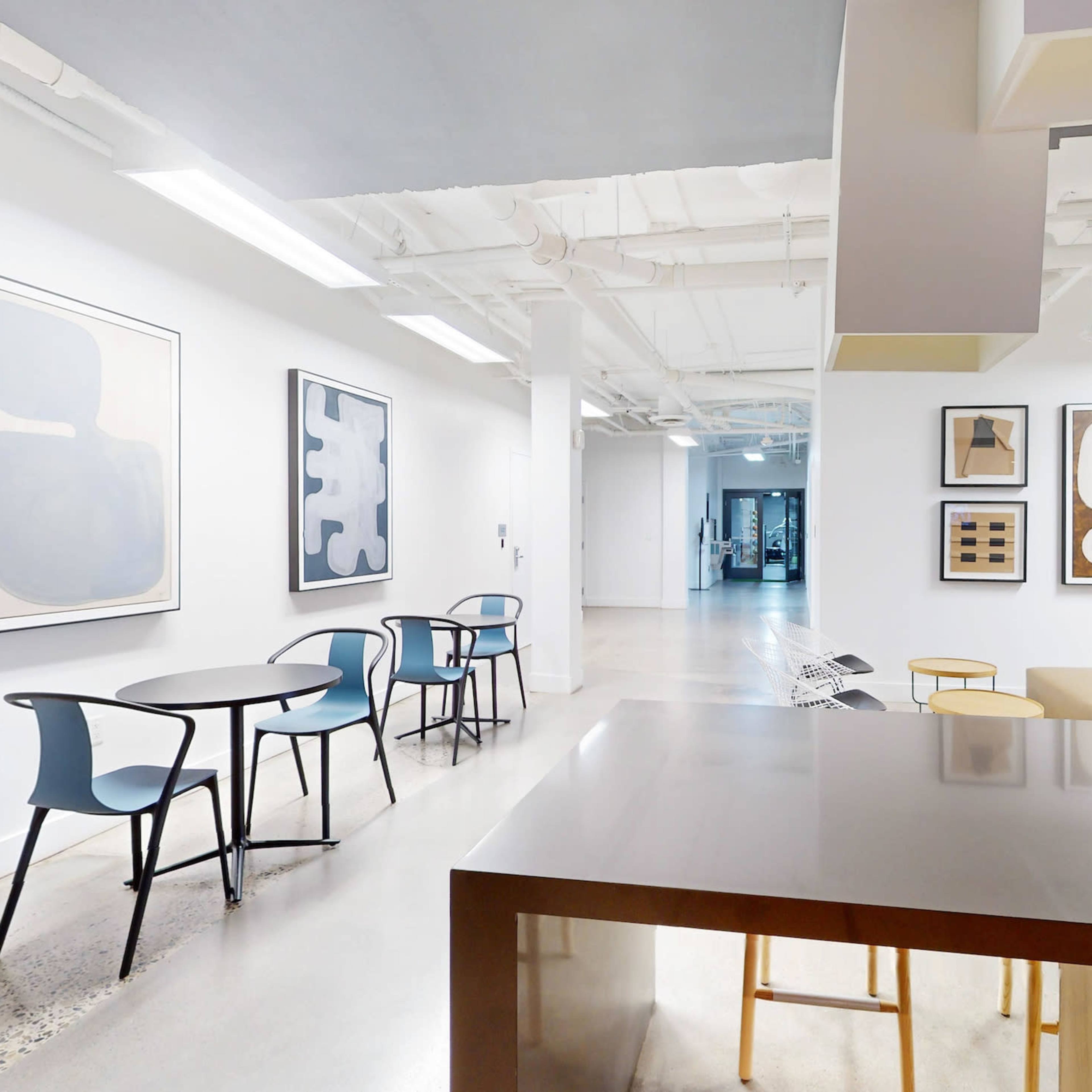 A modern interior space featuring a kitchenette, round tables with chairs, and several framed artworks on the white walls.
