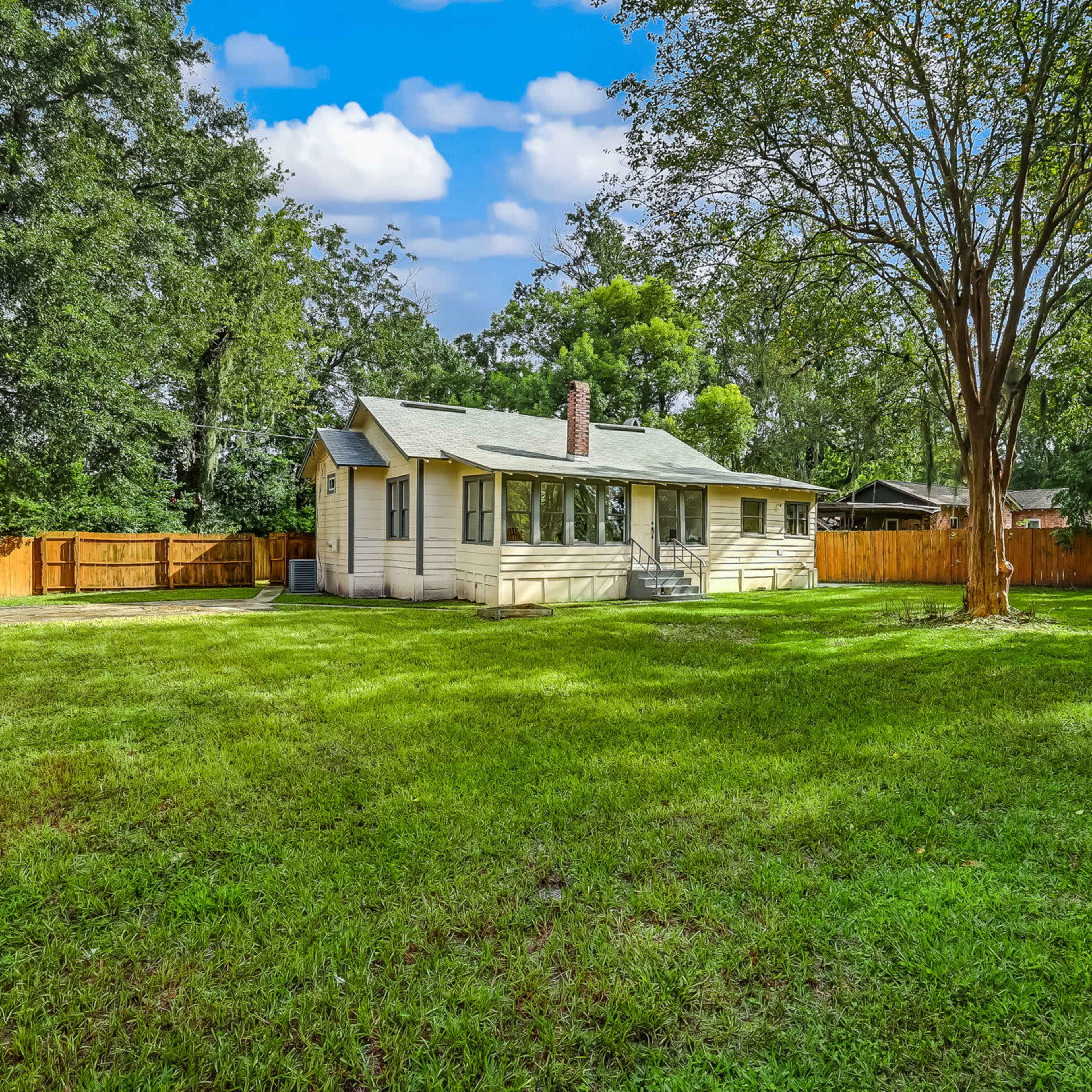 A single-story house with a front porch sits on a large grassy yard surrounded by trees and a wooden fence.