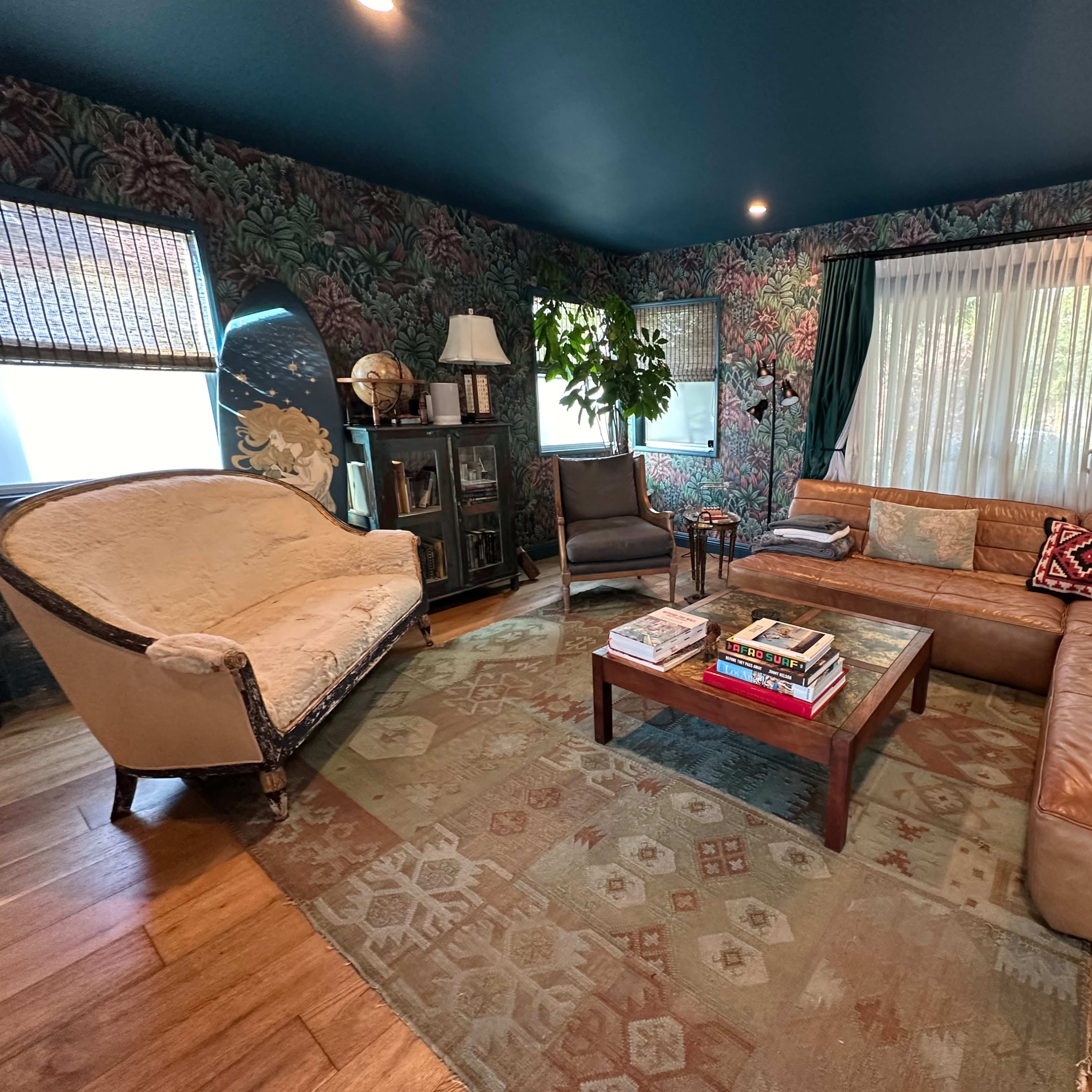 A cozy living room features a sectional leather sofa, a circular chair, a wooden coffee table stacked with books, and wallpaper adorned with floral designs.