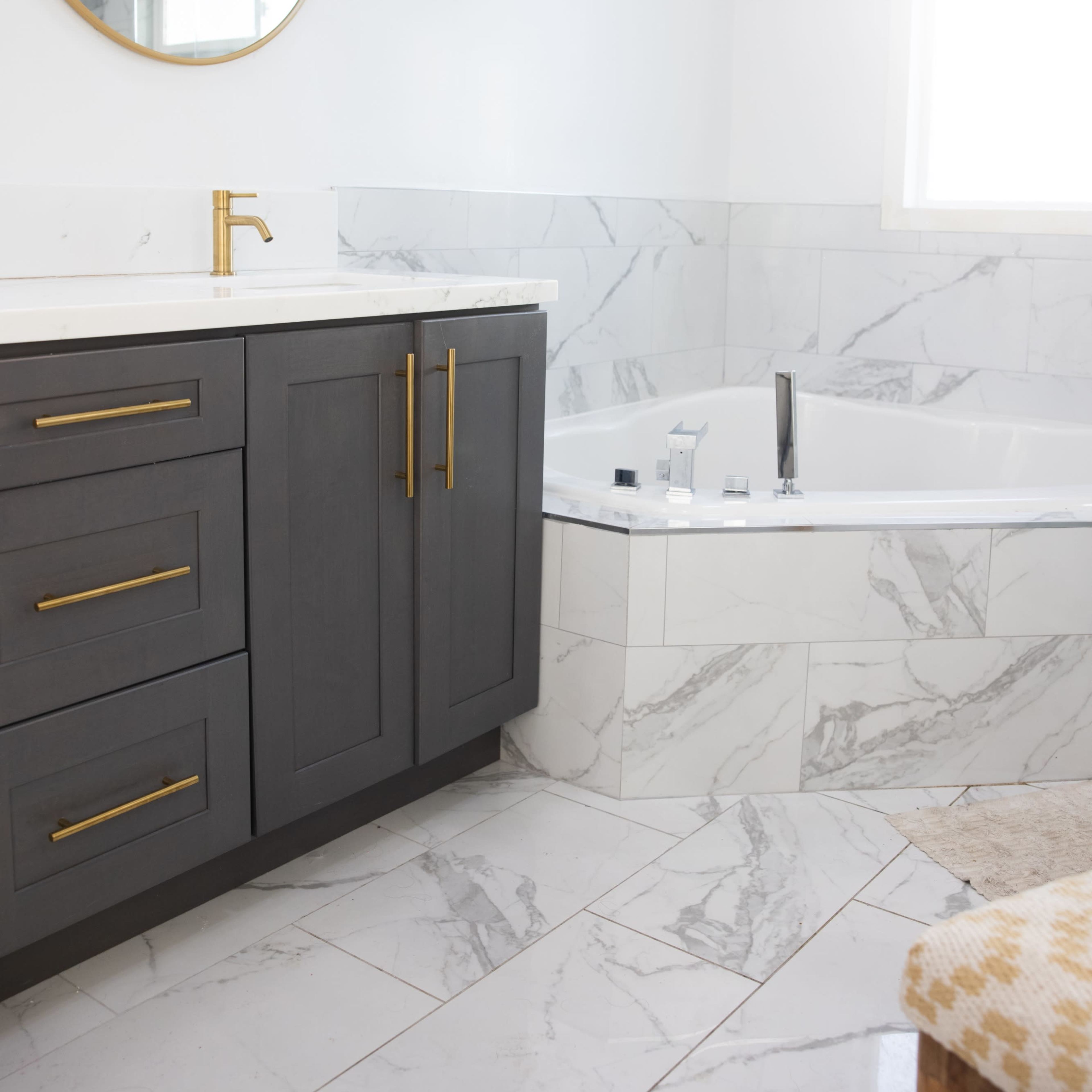 A modern bathroom features a dark gray vanity with gold hardware, a circular mirror, a white bathtub with marble surround, and marble flooring.