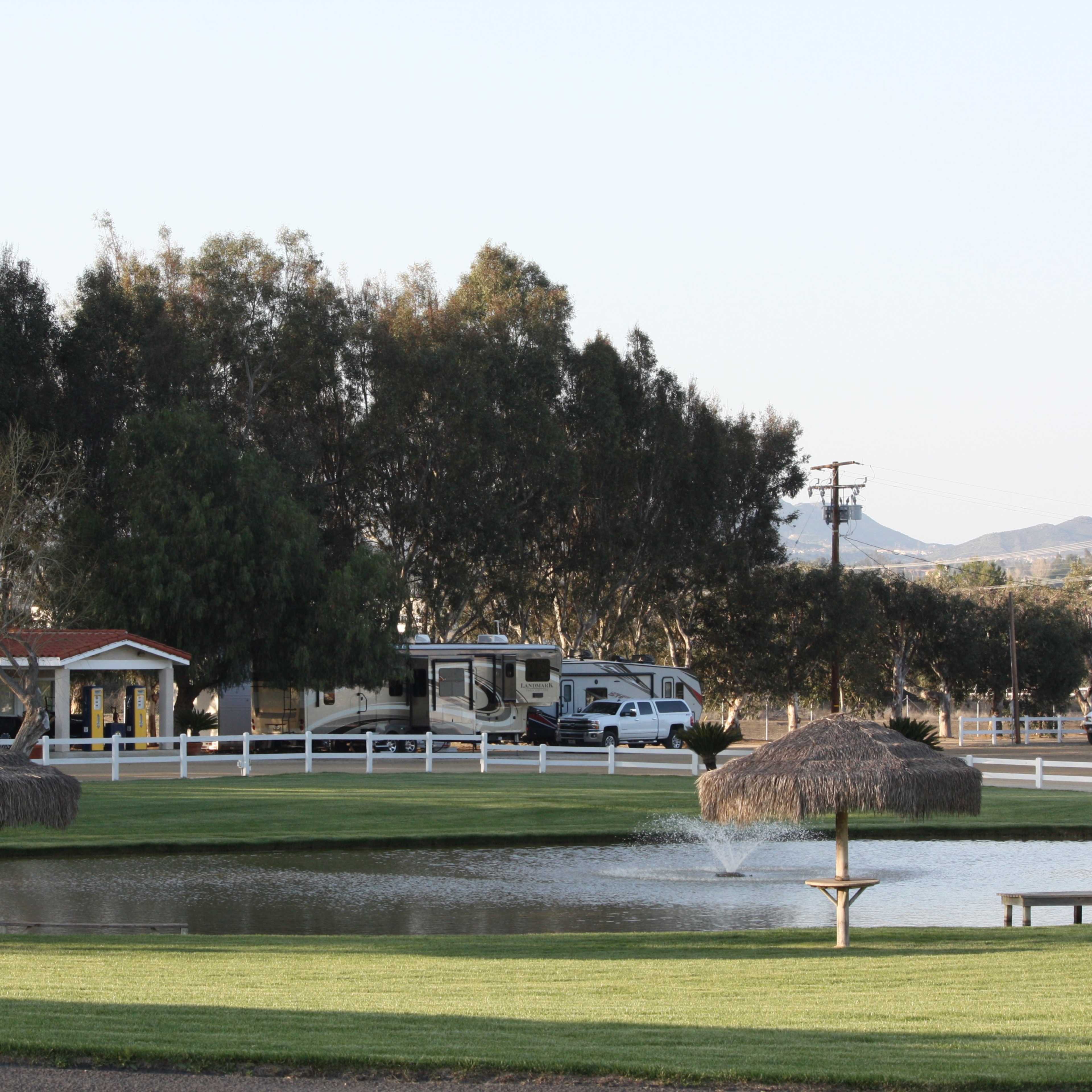 The scene shows a park with a pond, two thatched-roof structures, recreational vehicles in the background, and hills in the distance.