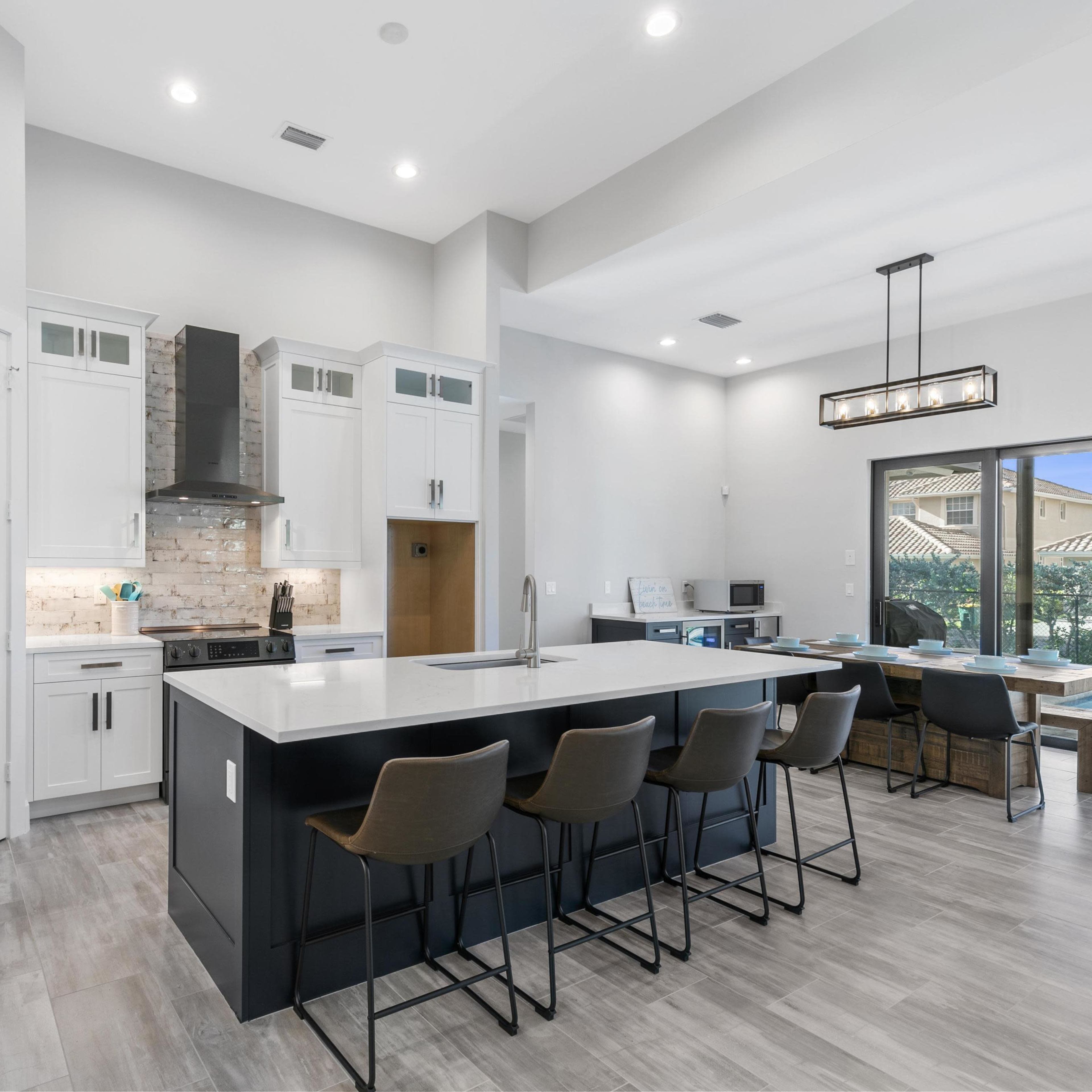 A modern kitchen with a large island, black bar stools, white cabinetry, and a view of an outdoor patio through sliding glass doors.