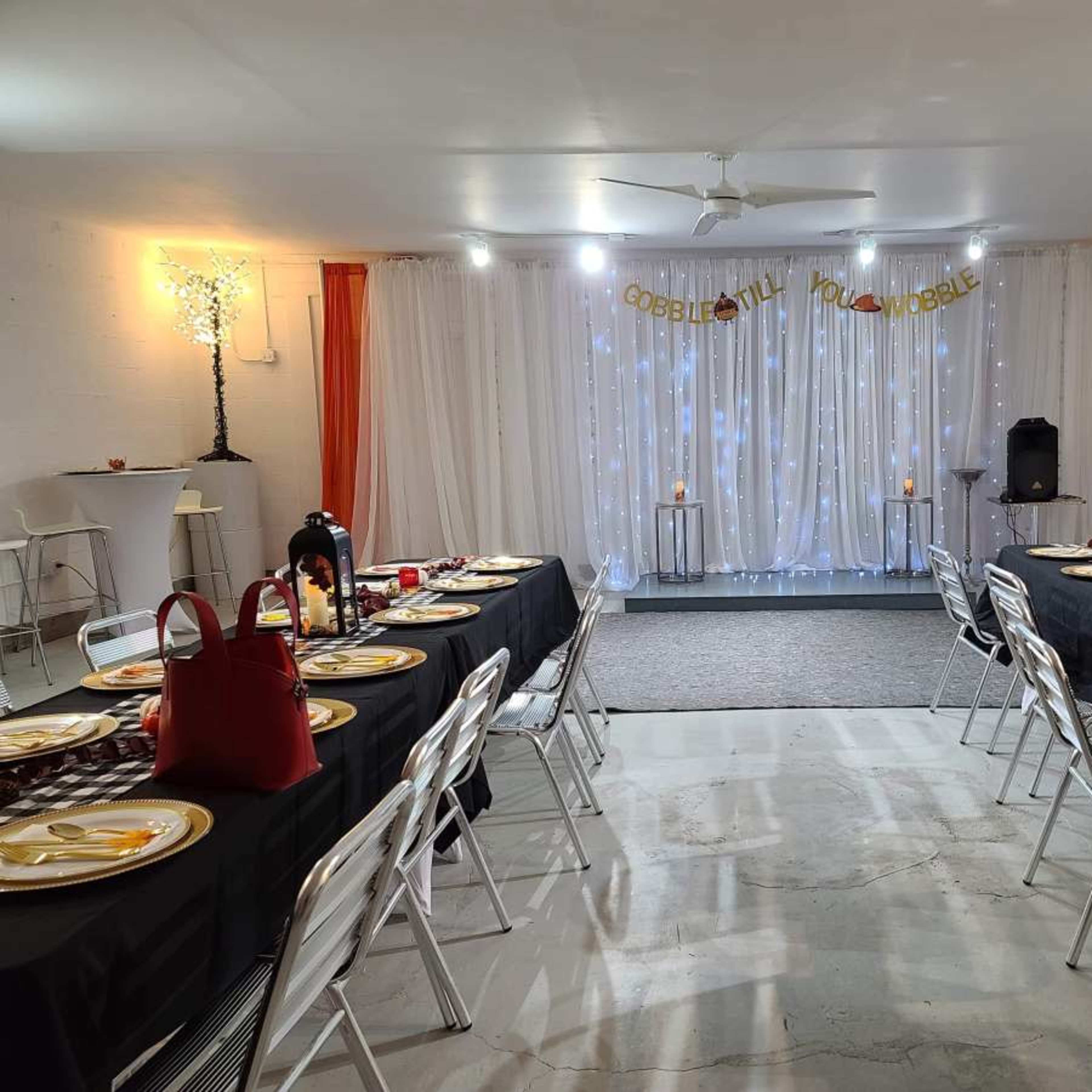 A dining area is set up with black tablecloths and gold plates, illuminated by festive lighting and decorative elements for a gathering.