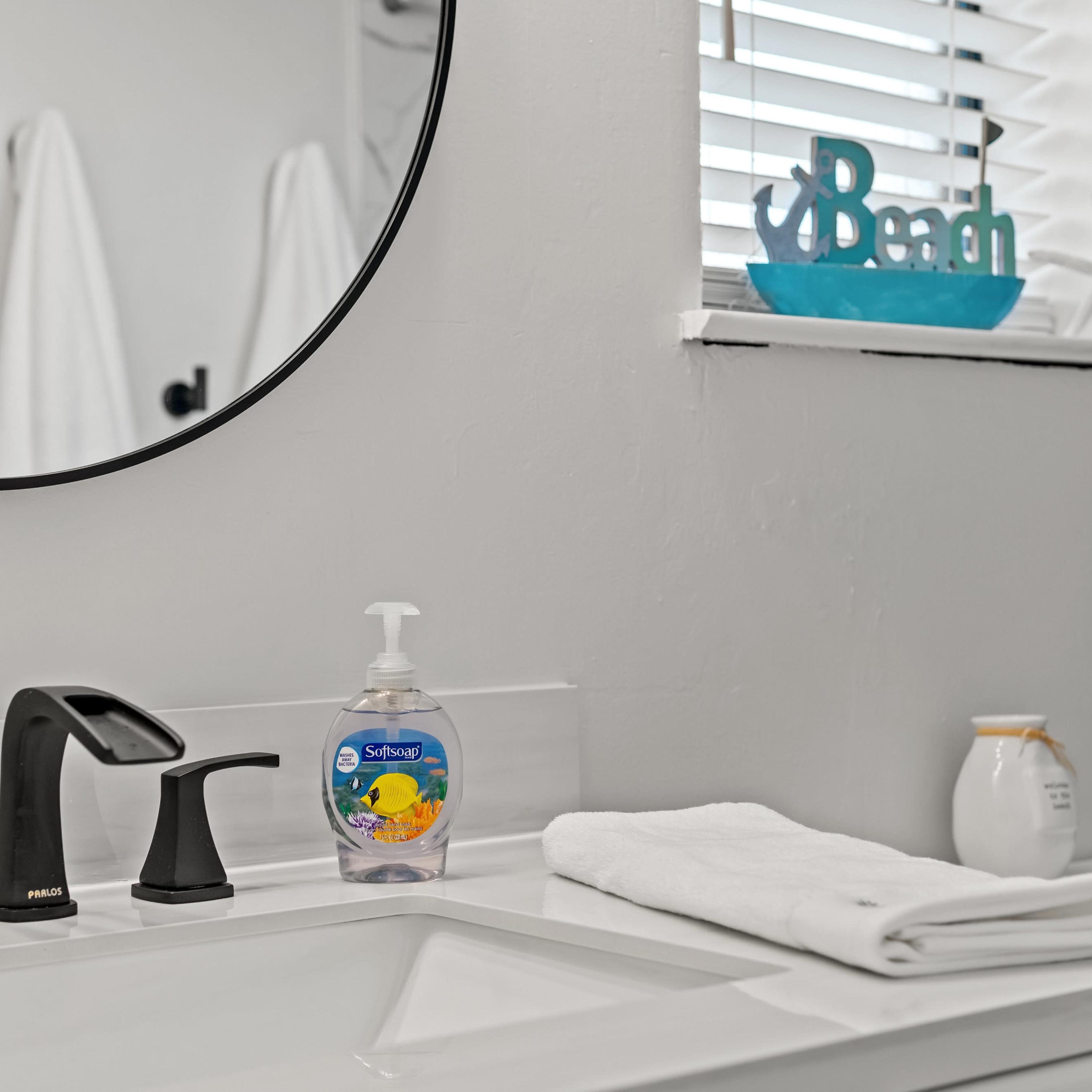 A bathroom sink area features a black faucet, a bottle of hand soap, a rolled white towel, and a small decorative item labeled "Beach" near a mirror.