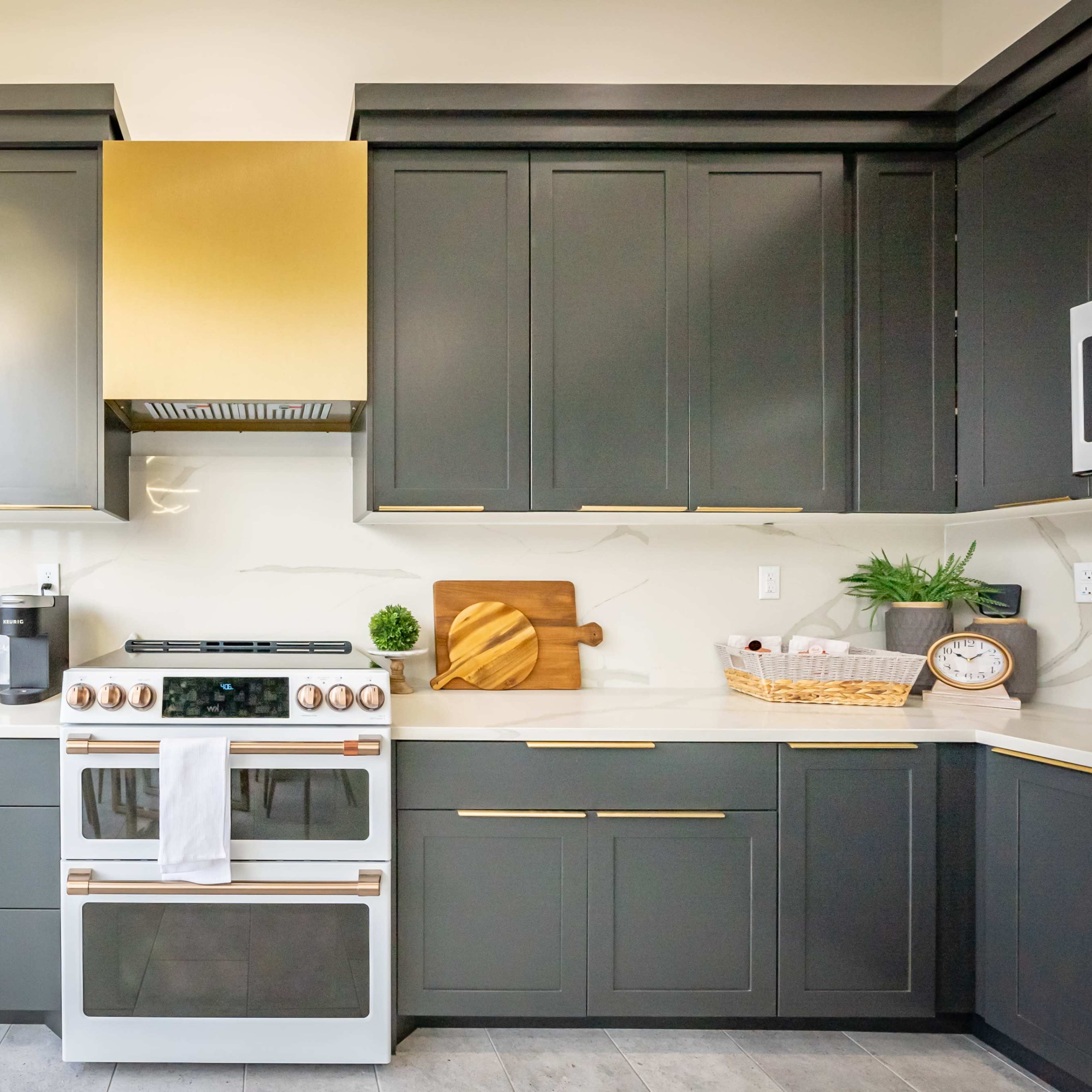 A modern kitchen features dark cabinetry, a gold-accented range hood, a white stove, and various kitchen appliances arranged on the countertops.