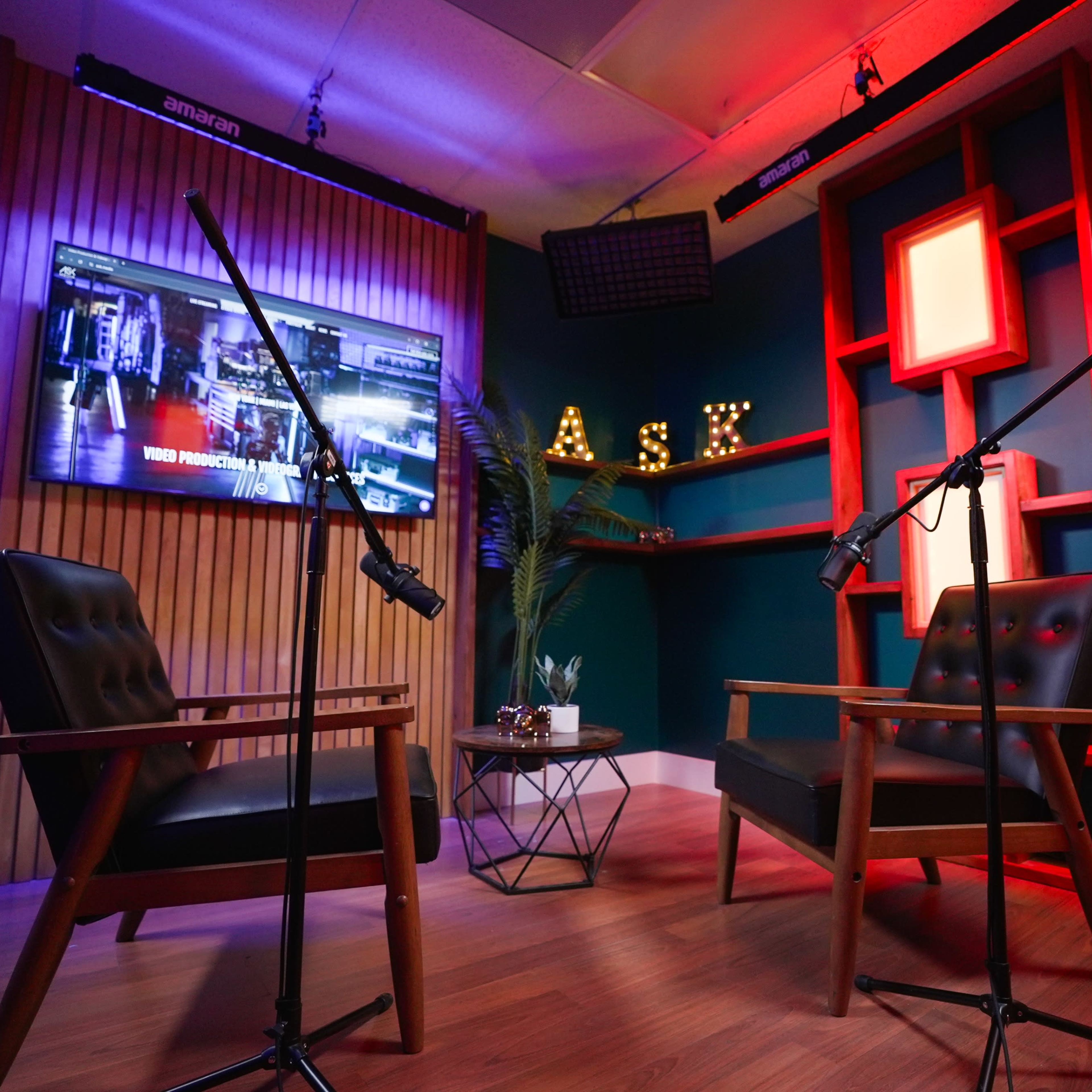 A small studio setup with two chairs, each equipped with a microphone on a stand, and a brightly lit background featuring wooden panels and a television screen.