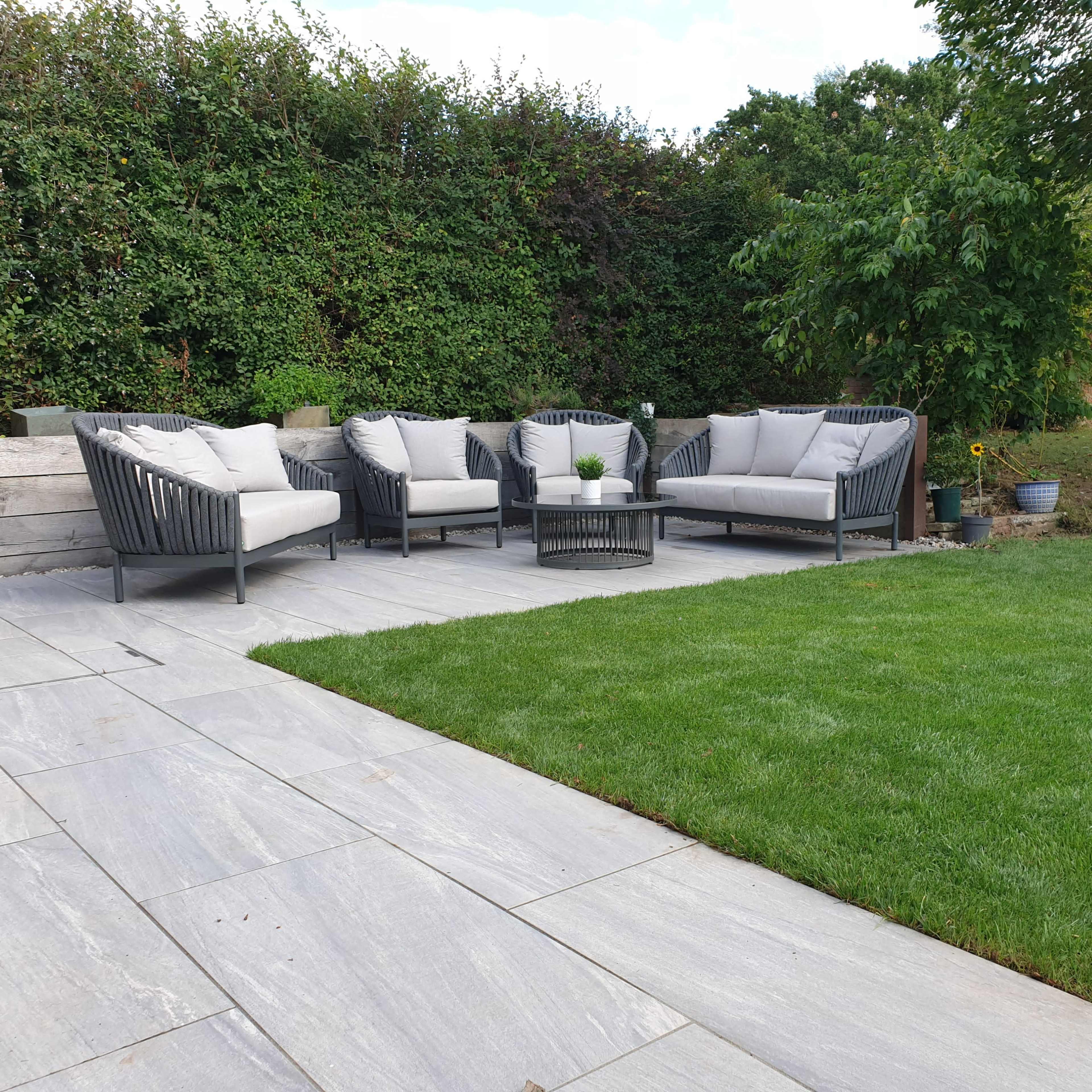 A patio area with gray stone flooring, a set of four wicker chairs with cushions, and a small table, surrounded by green grass and shrubs.