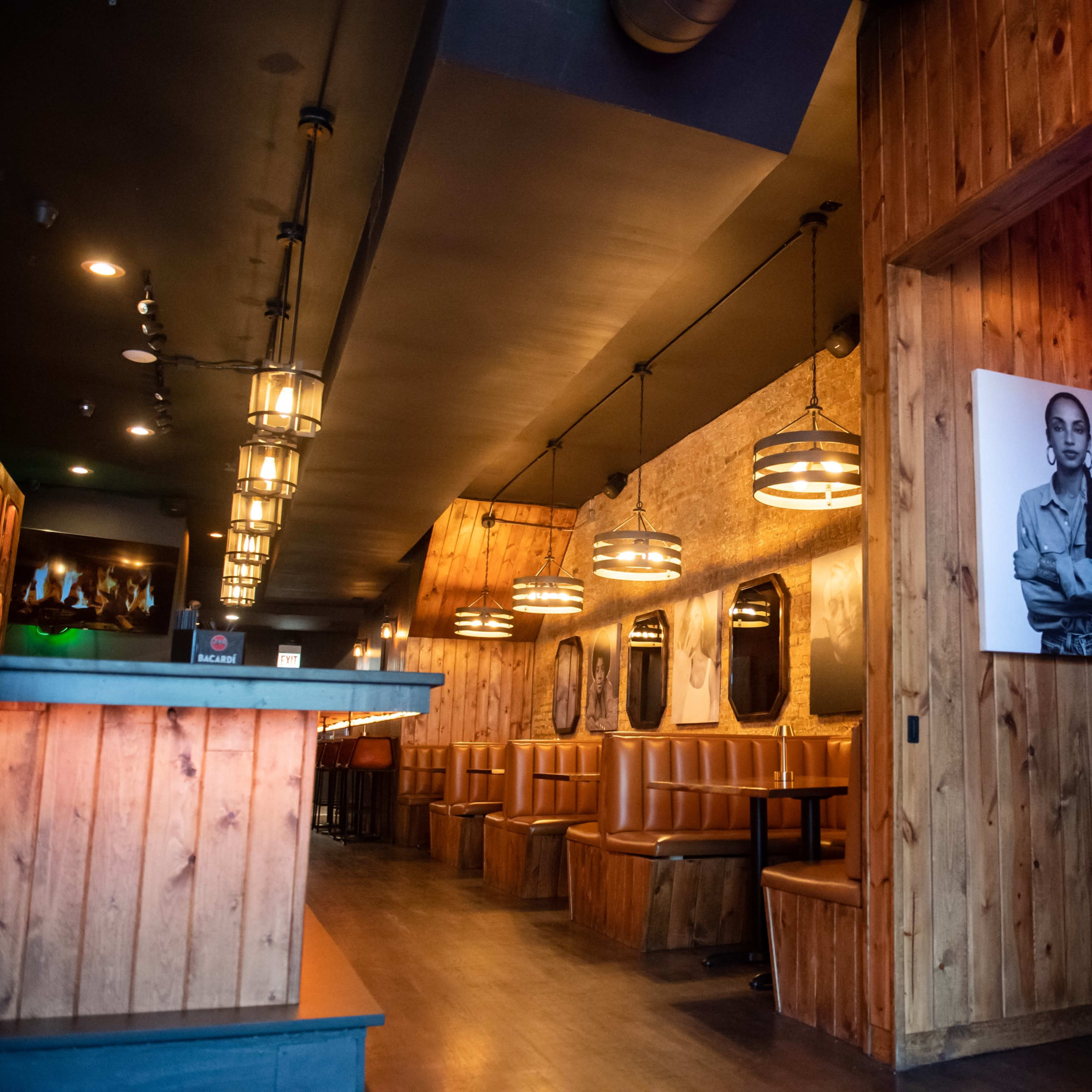 The interior of a restaurant with wooden paneling, booths along the walls, pendant lights hanging from the ceiling, and a neon sign that reads "Save Water Drink Champagne."