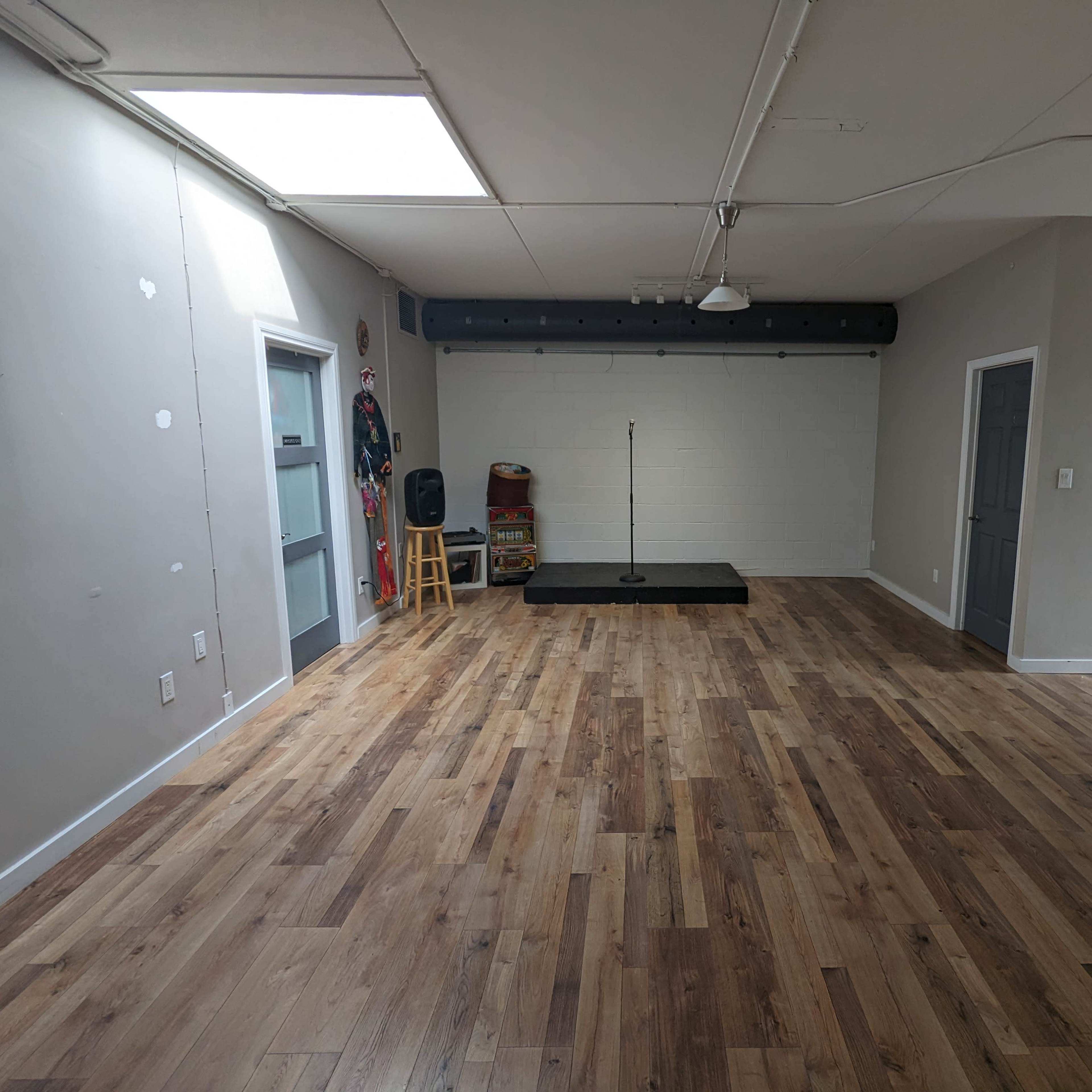 An empty room with wooden flooring, a stage at one end, a microphone stand, and walls painted in a light color.