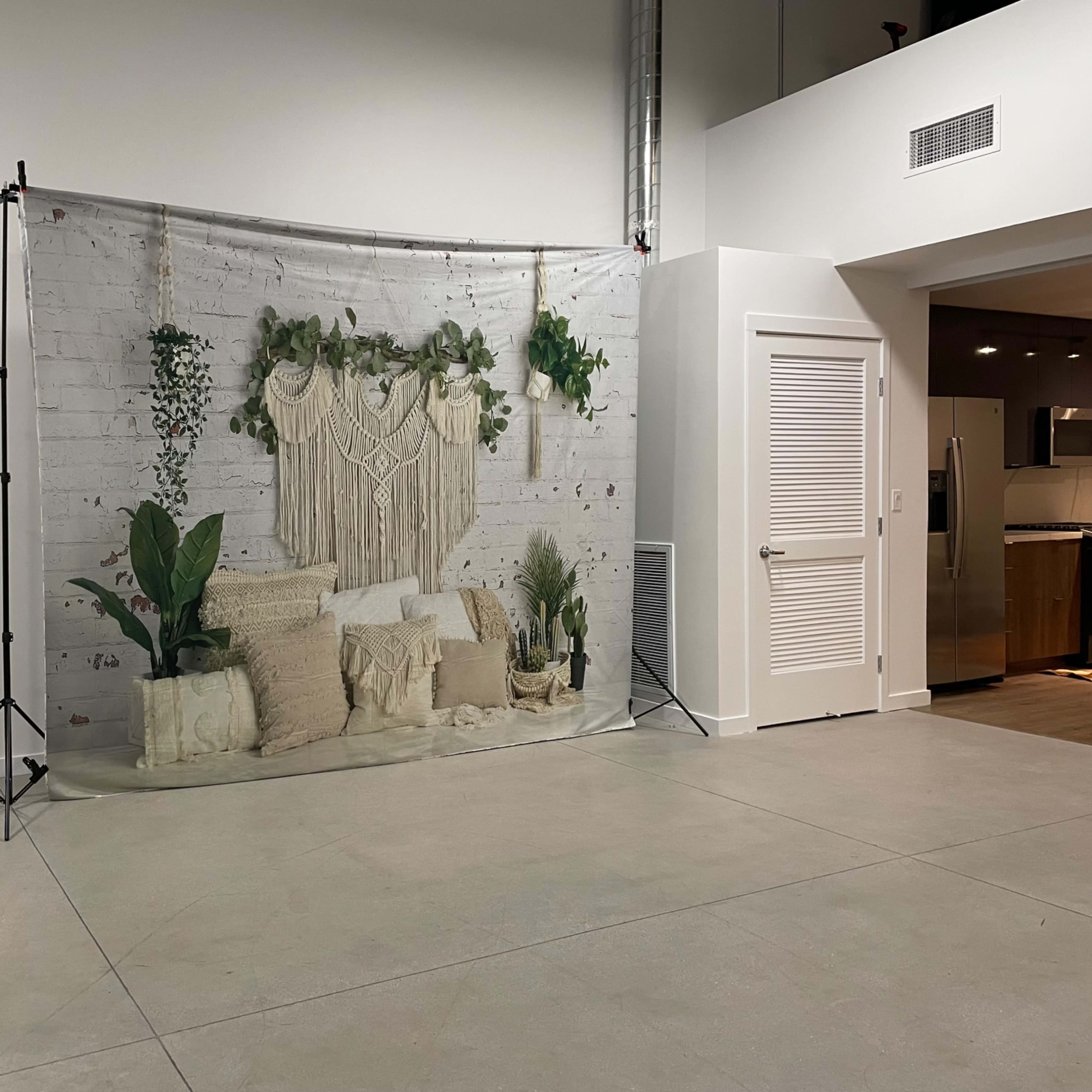 A decorative backdrop featuring a macramé wall hanging and various plants is positioned in a spacious, minimalist room with concrete flooring.
