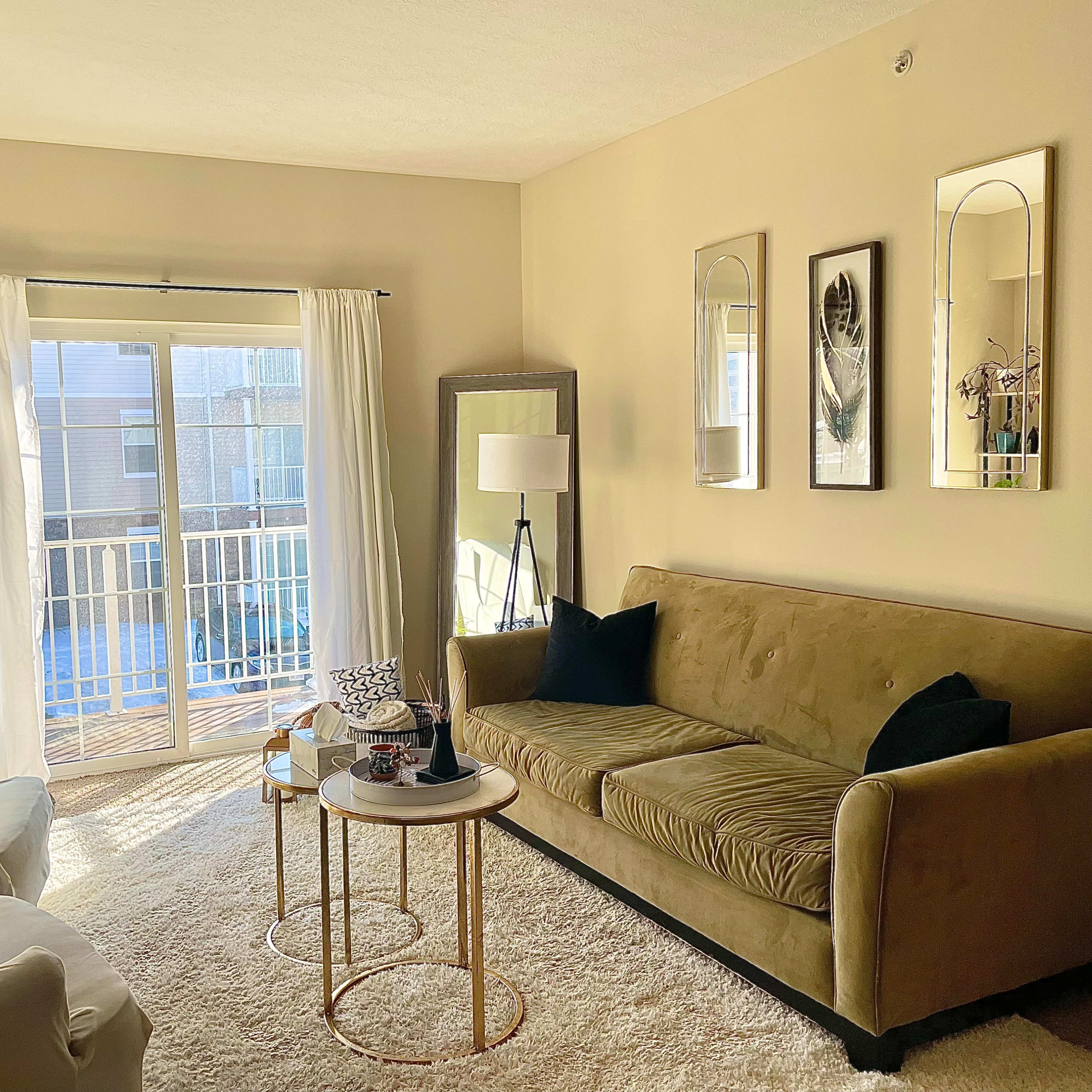A cozy living room with a green sofa, two side tables, and large windows revealing a balcony.