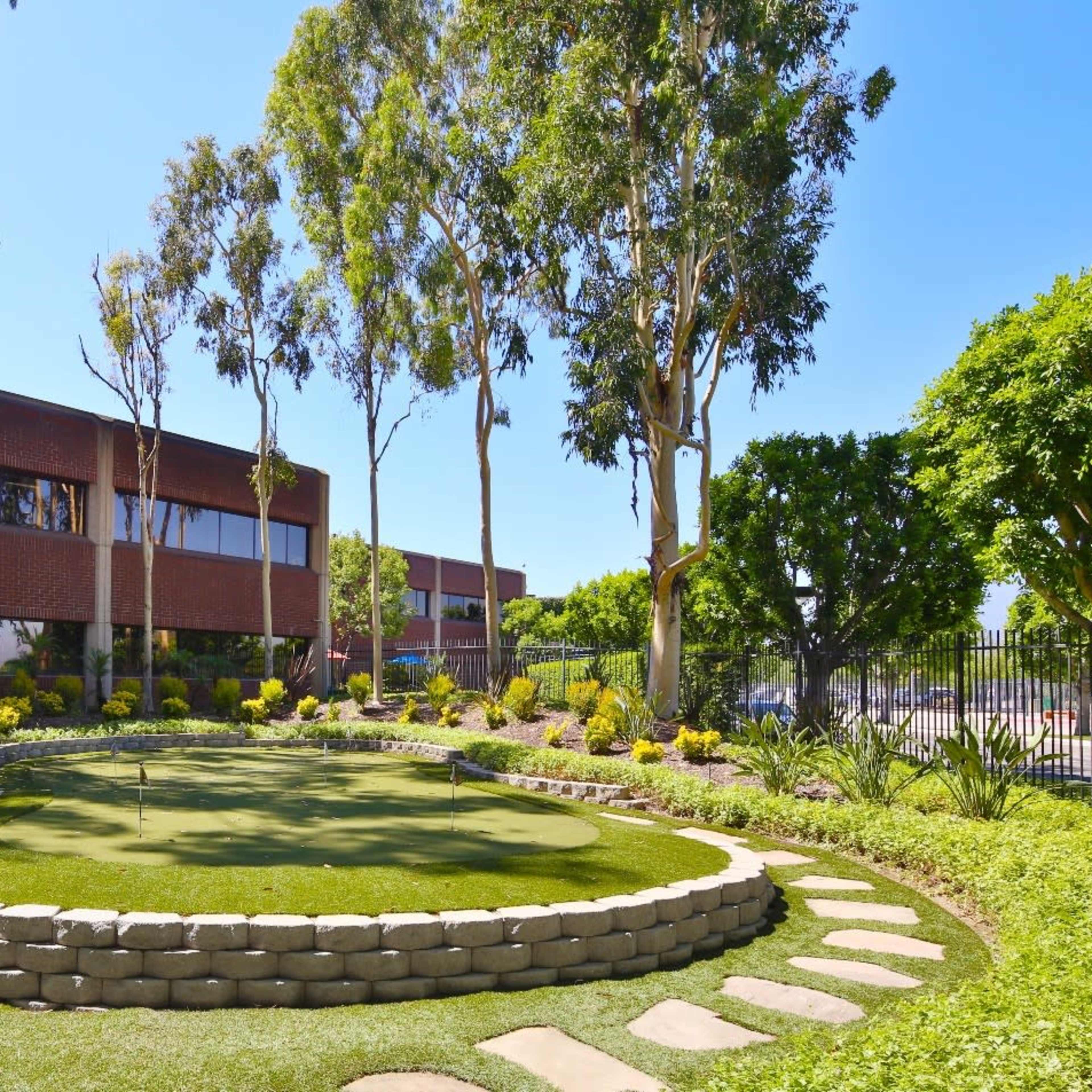 A landscaped garden with a small putting green bordered by stone, palm trees, and a brick building in the background.