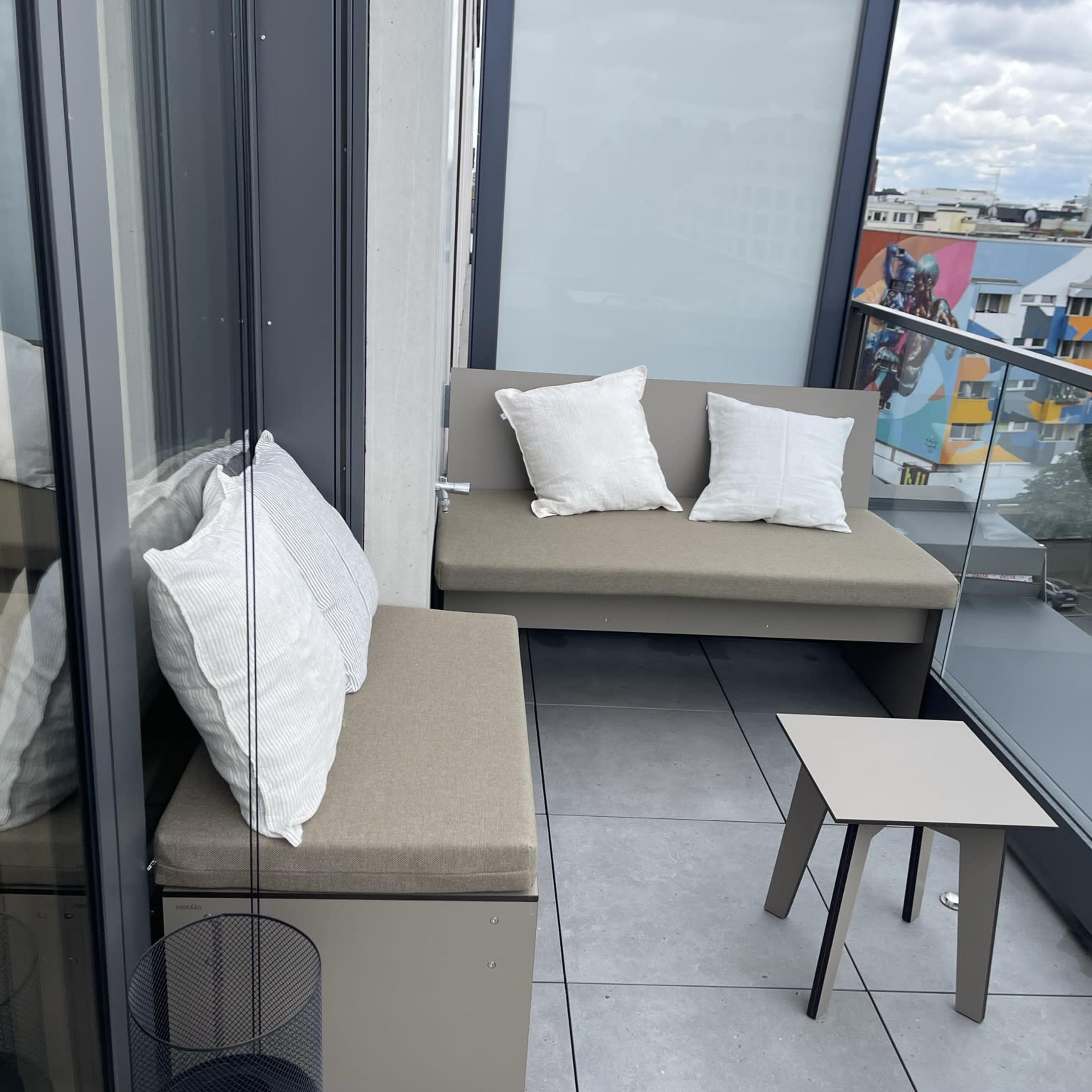 A balcony features a small seating area with a bench and two cushions, a side table, and a decorative planter.