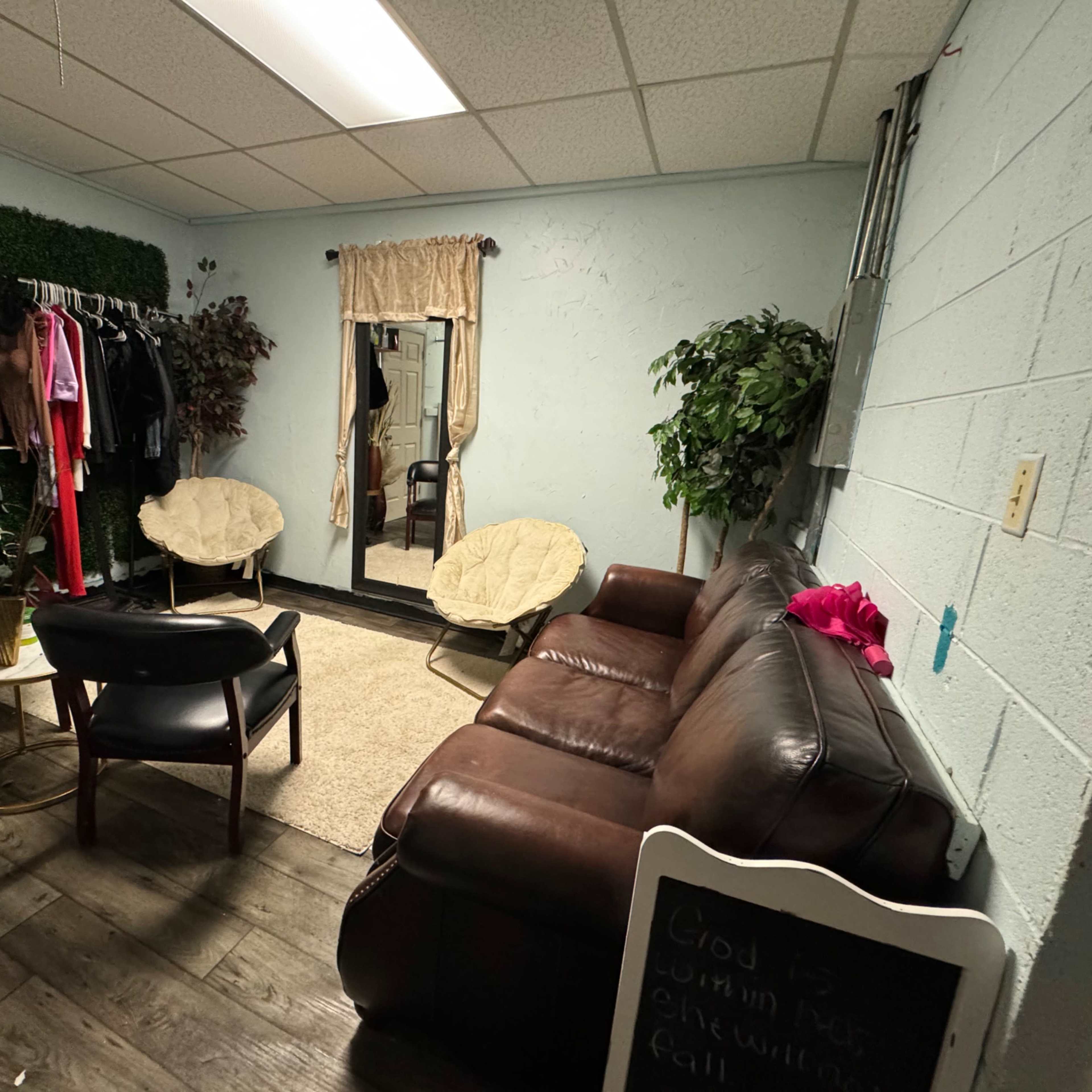 A small lounge area with a brown leather sofa, two chairs, a mirror, and clothes displayed on racks against a green wall.