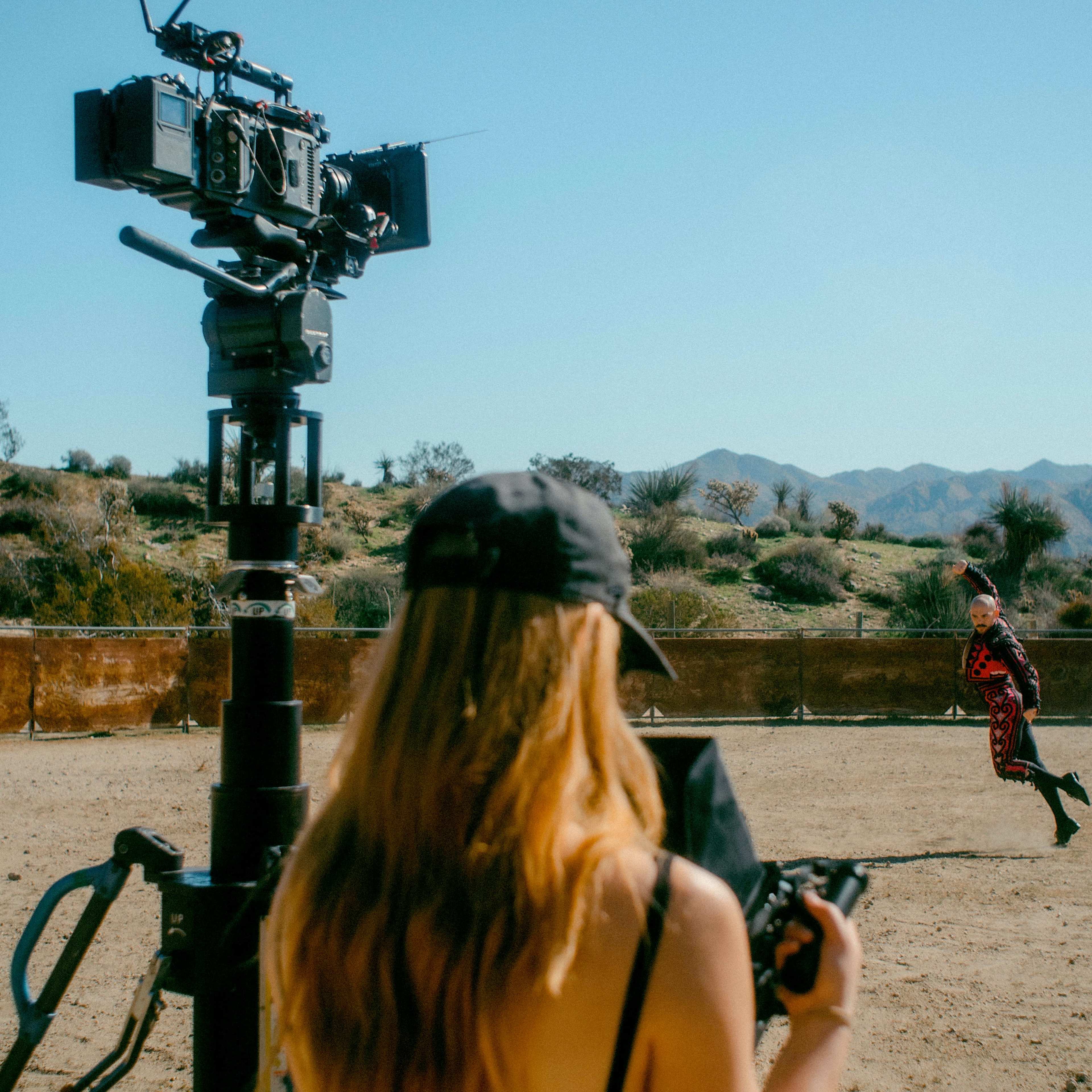 A camera operator captures a performer in a desert landscape as a film crew sets up equipment nearby.