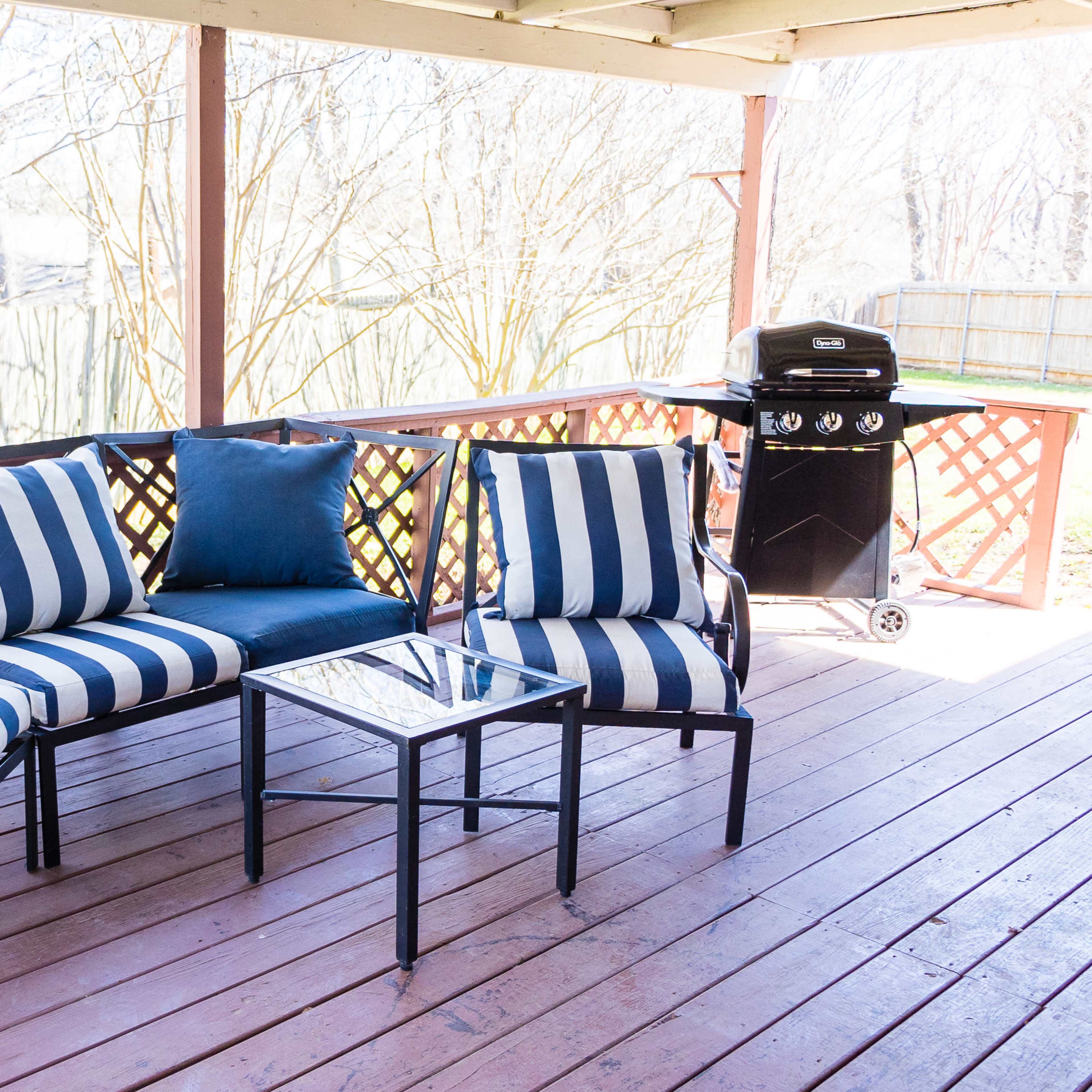 A patio area features a striped seating arrangement and a glass-top table next to a grill under a wooden pergola.