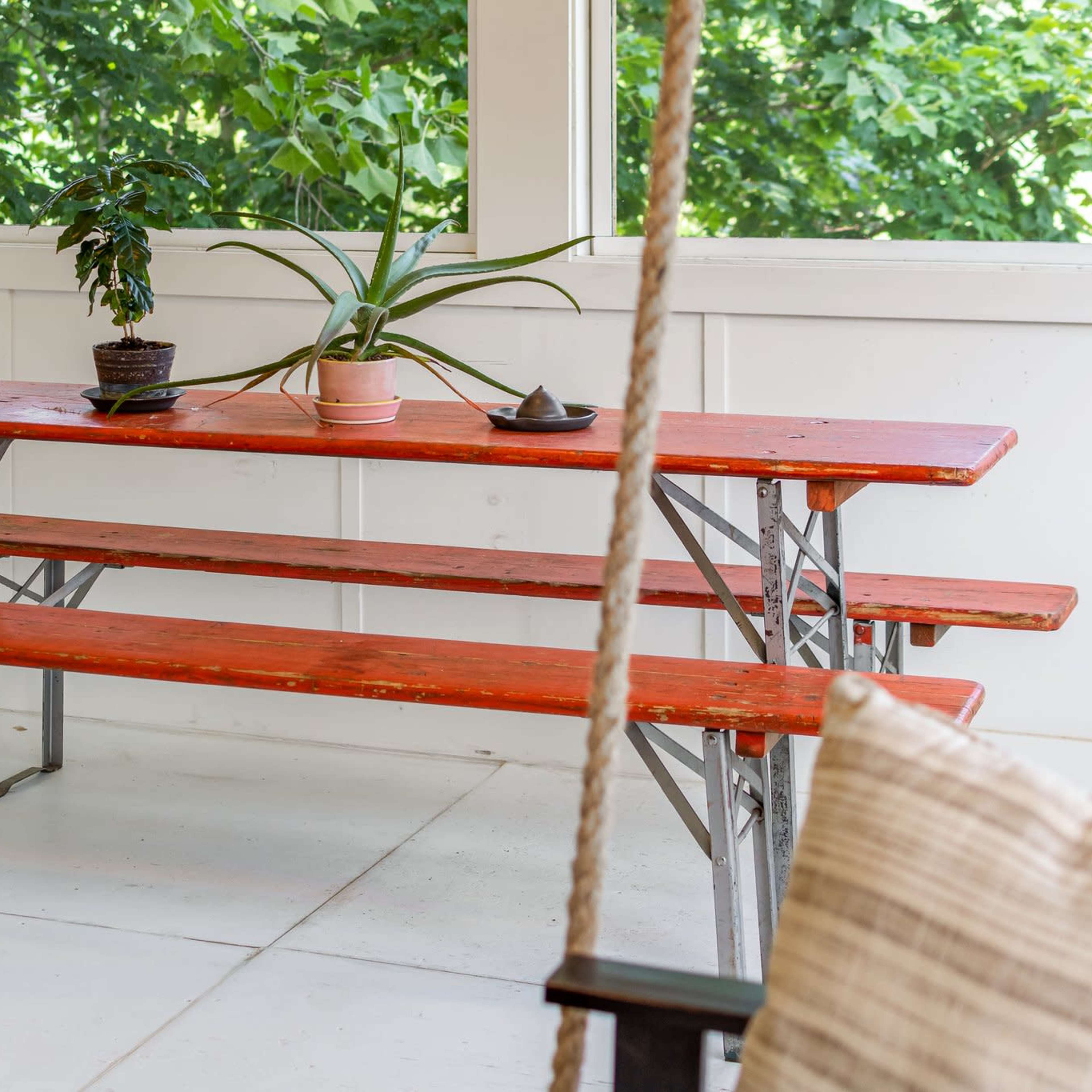 A wooden picnic table with two benches and potted plants sits in a brightly lit room with large windows.
