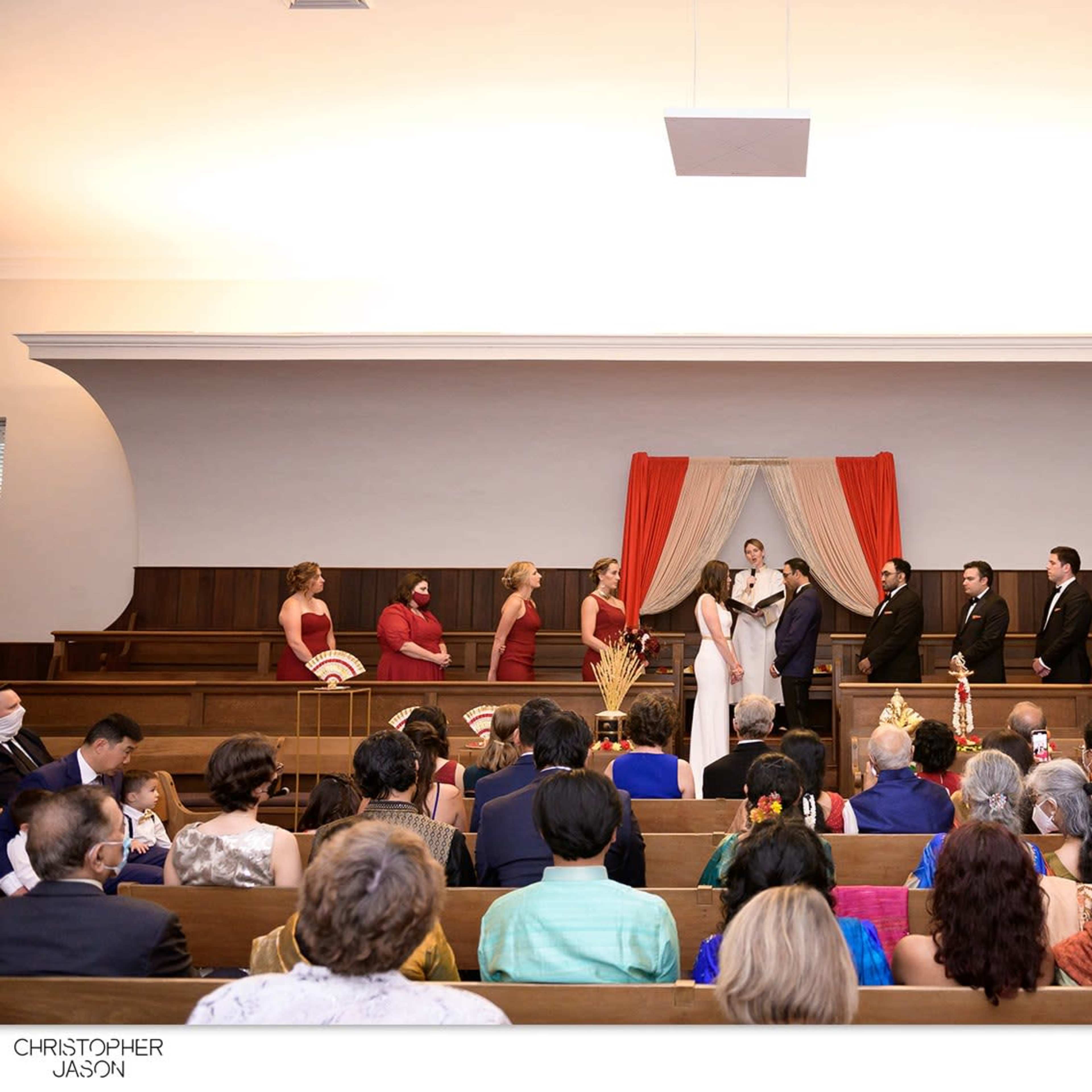 A wedding ceremony is taking place in a venue with a decorated altar, featuring a couple in the center and guests seated in wooden pews.
