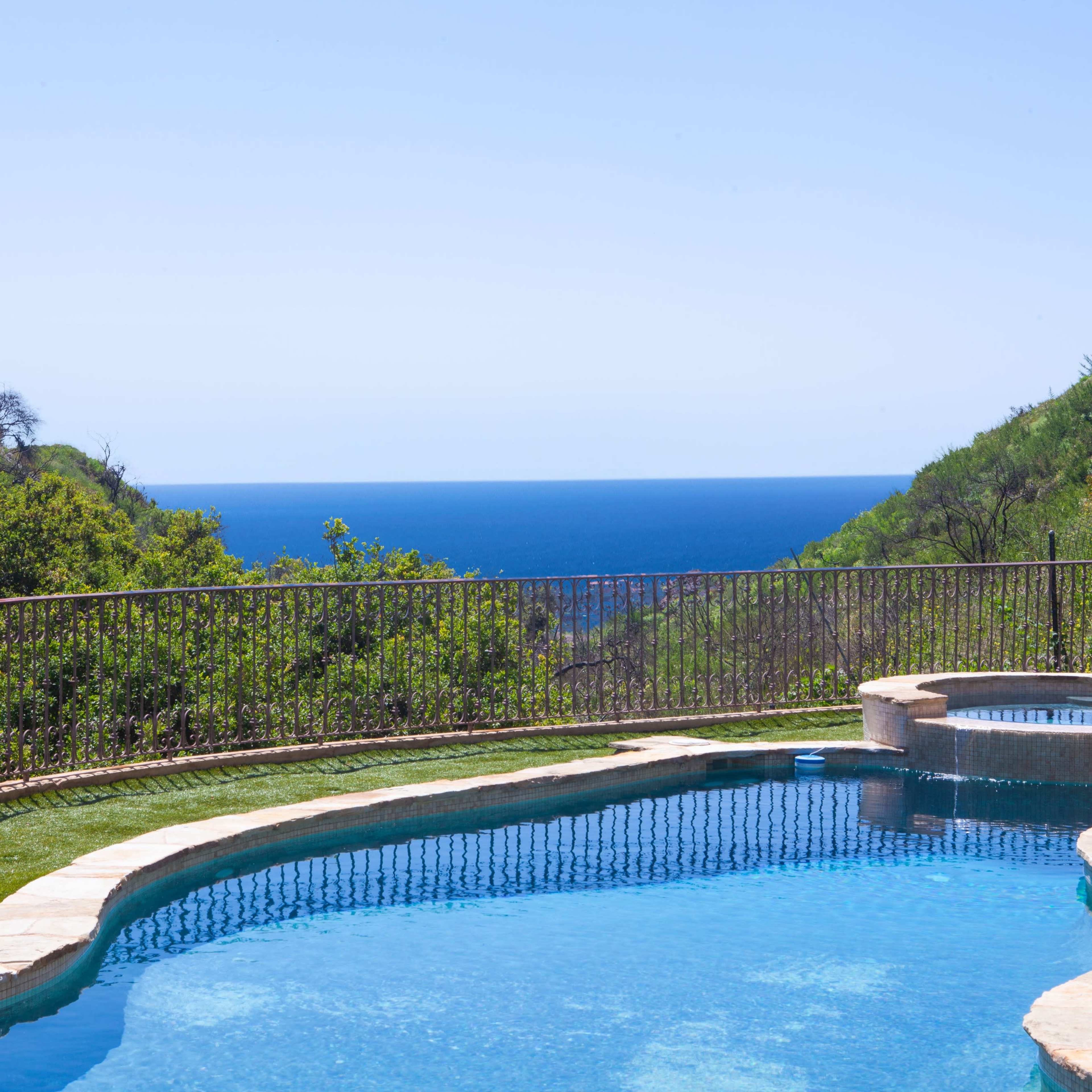 A swimming pool with a spa overlooks a blue ocean surrounded by green hills.