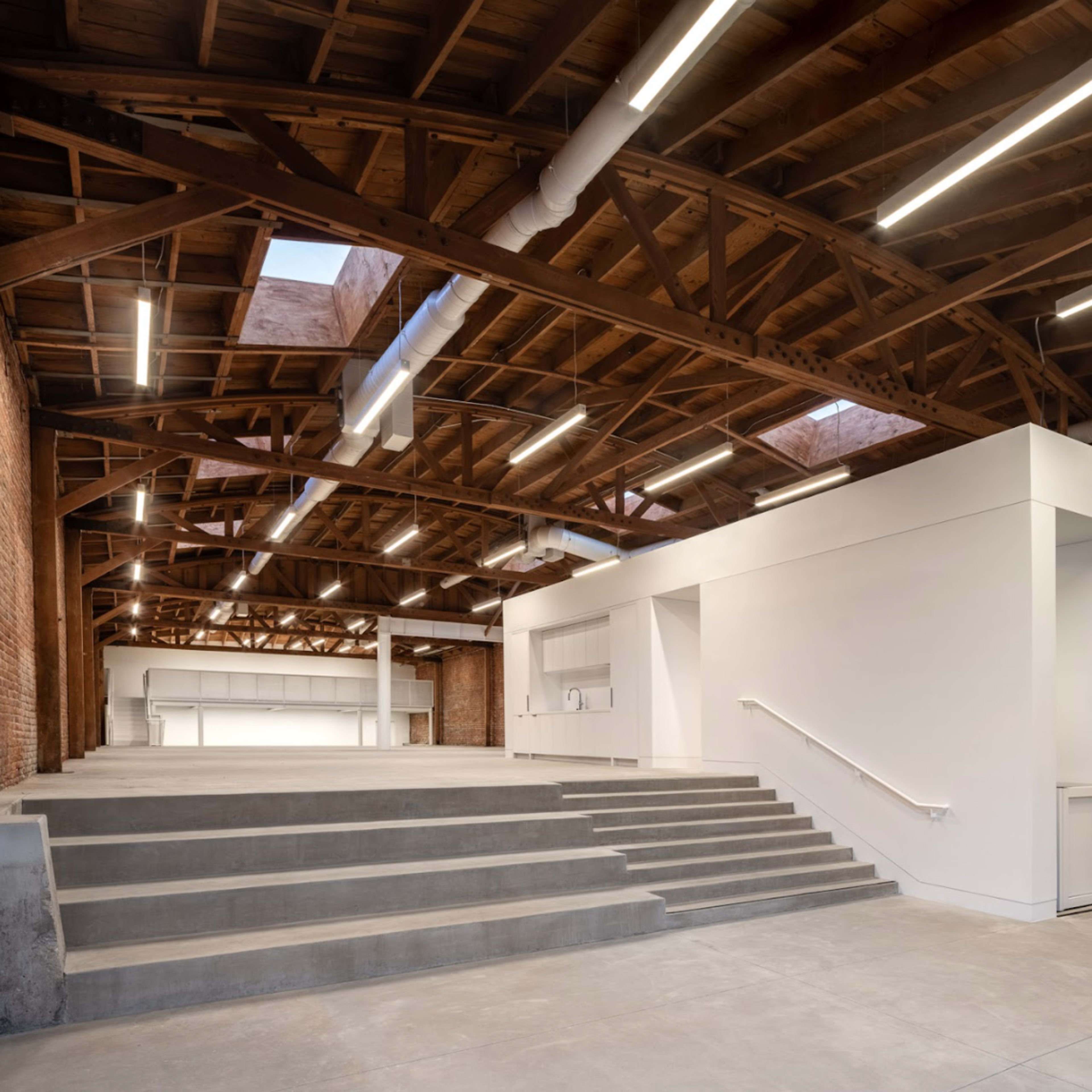 An industrial-style interior with exposed brick walls, wooden beams, and a staircase leading to a raised platform in a spacious room.
