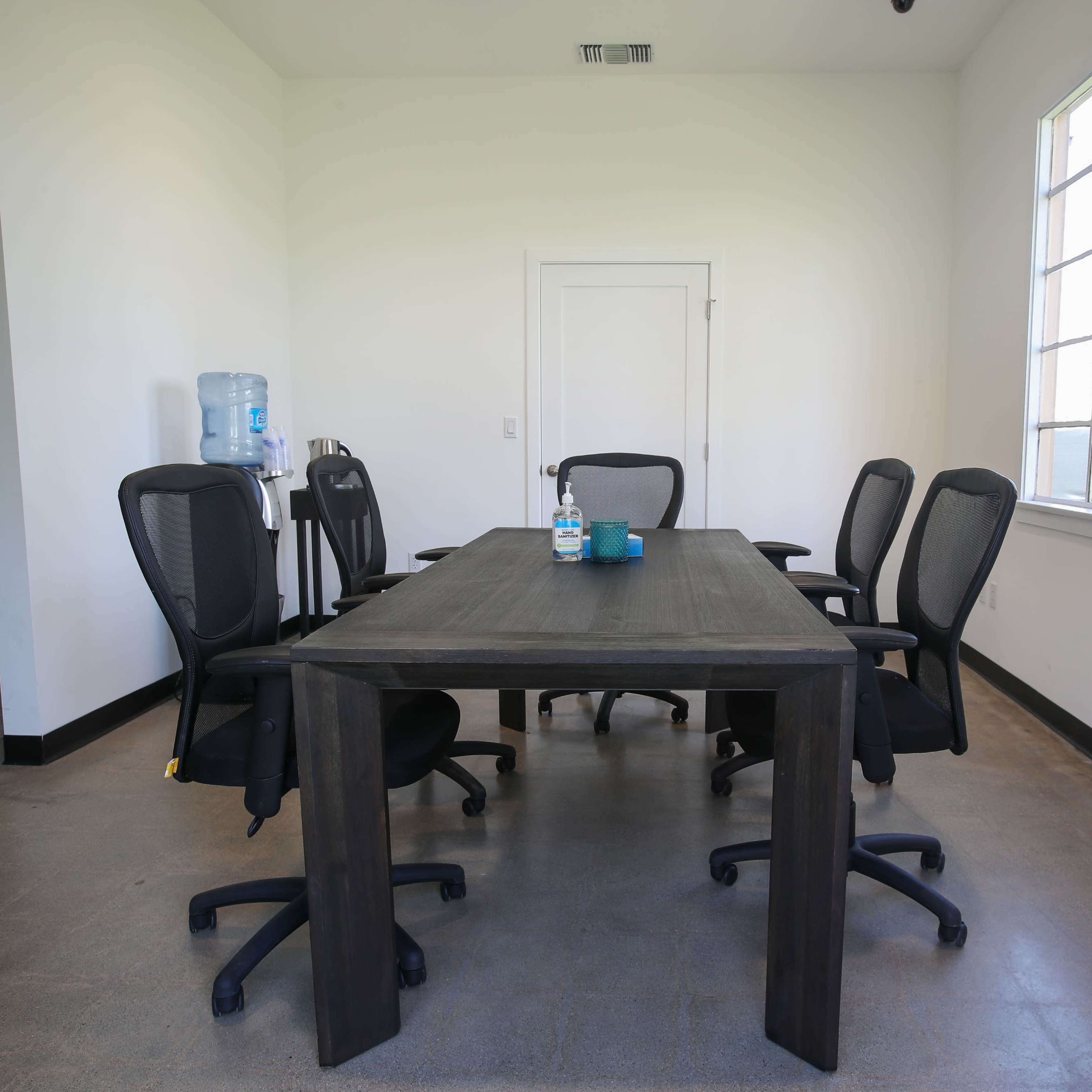 A conference room with a large wooden table surrounded by black office chairs, a water dispenser in the corner, and a window letting in natural light.