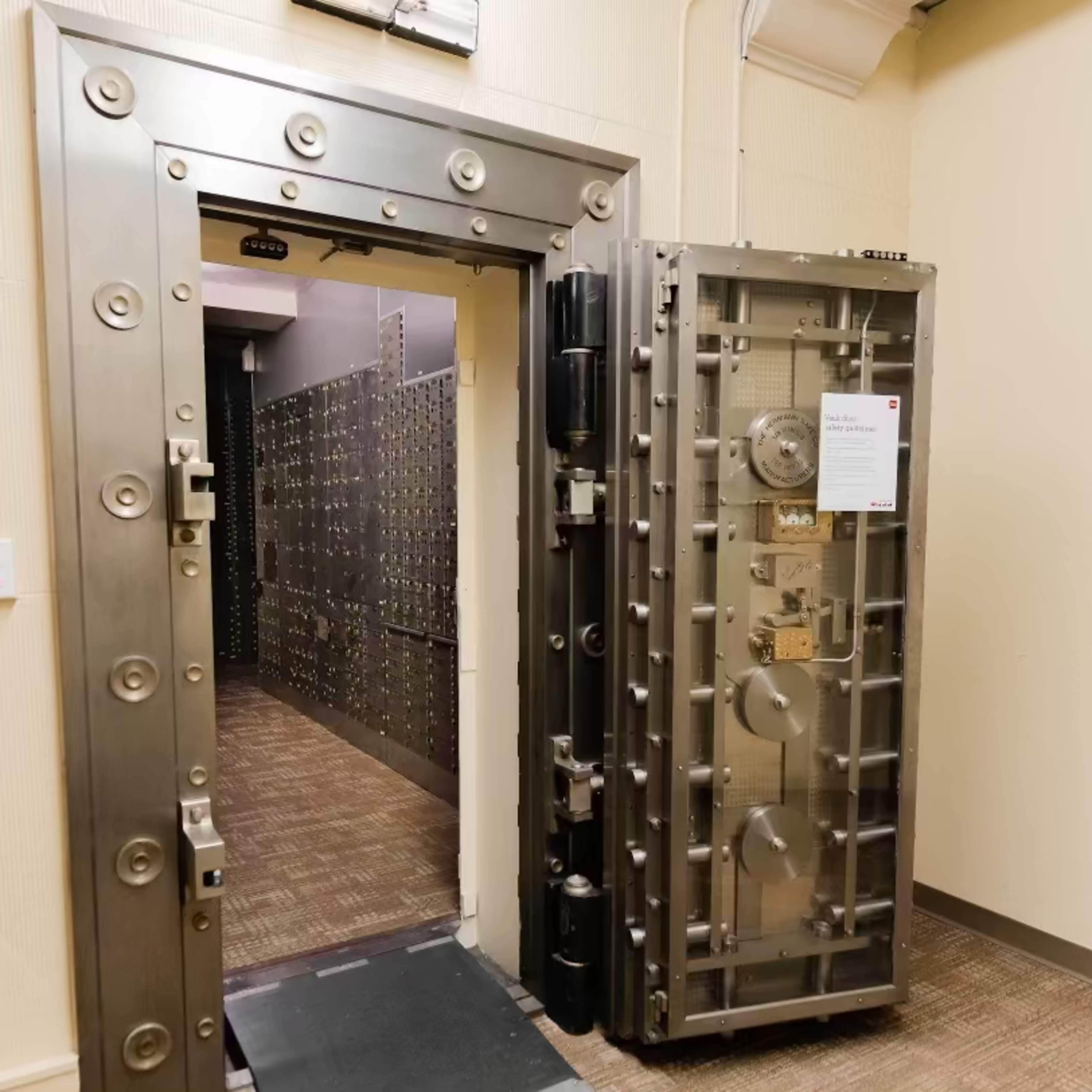 A large, metallic vault door stands open, revealing a corridor lined with smaller safe deposit boxes.