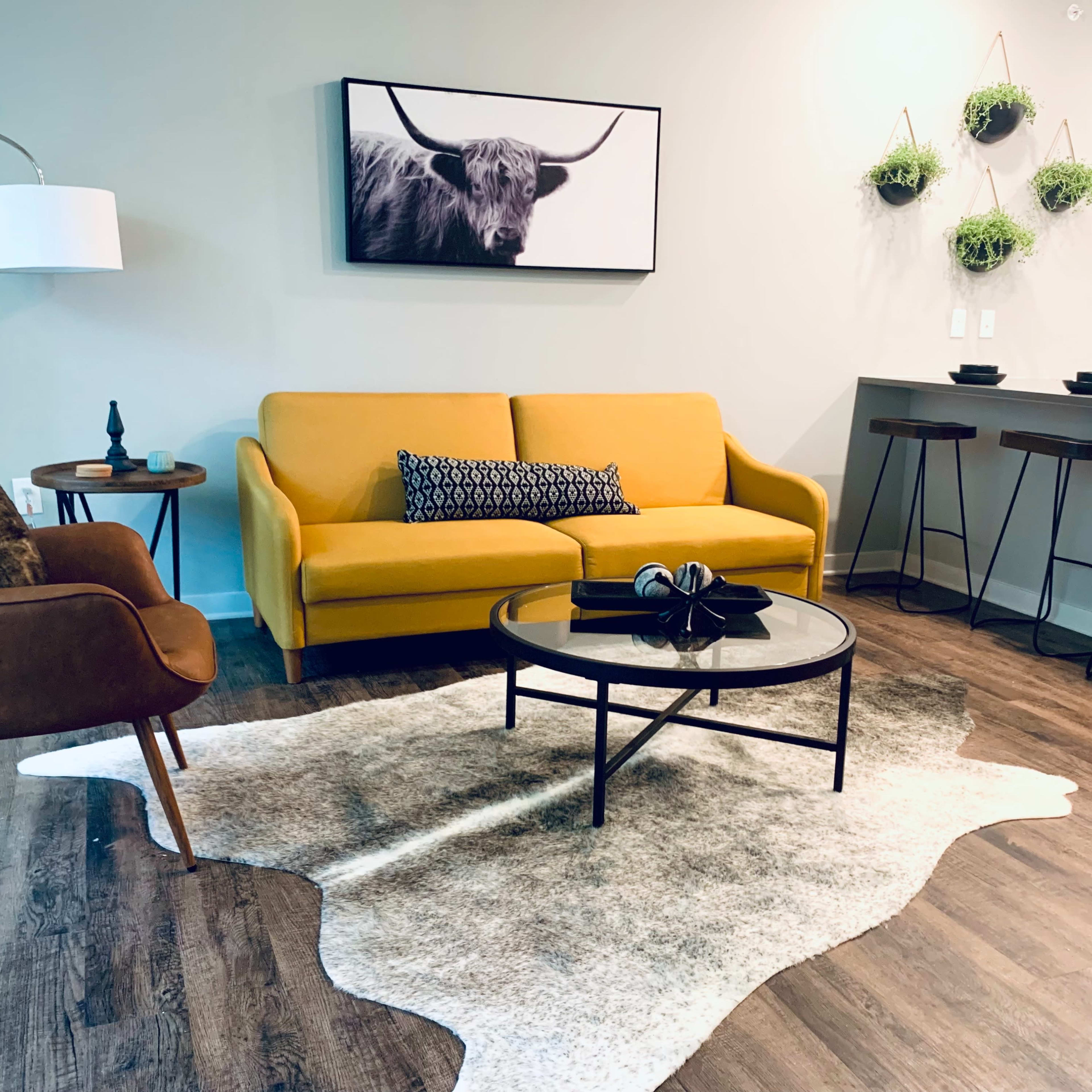 The living area features a yellow sofa, an armchair, a coffee table, and a wall-mounted artwork of a bull above a modern kitchenette with bar stools.