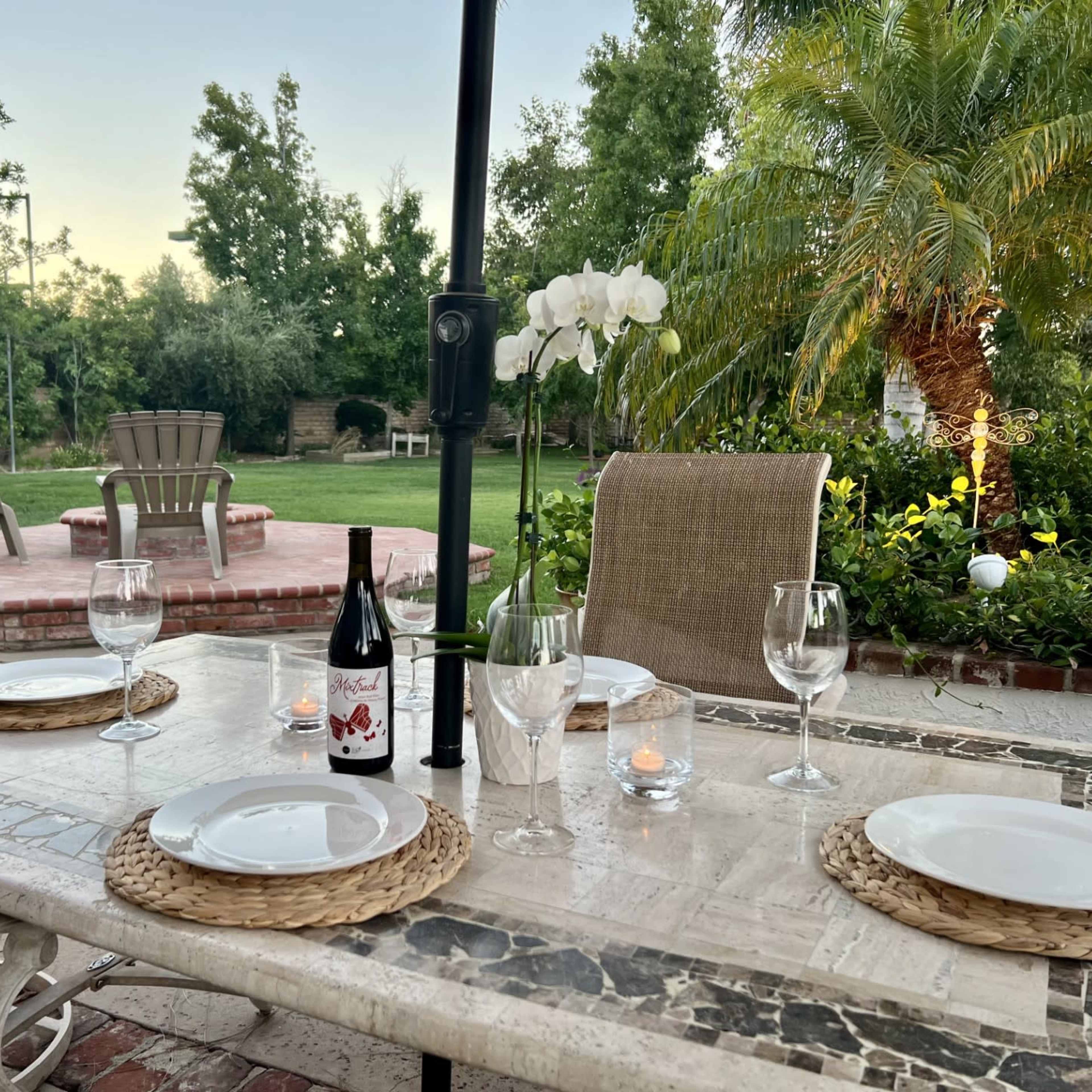 A neatly arranged outdoor dining table is set for four, featuring plates, glasses, a wine bottle, and candles, with a lush garden backdrop.