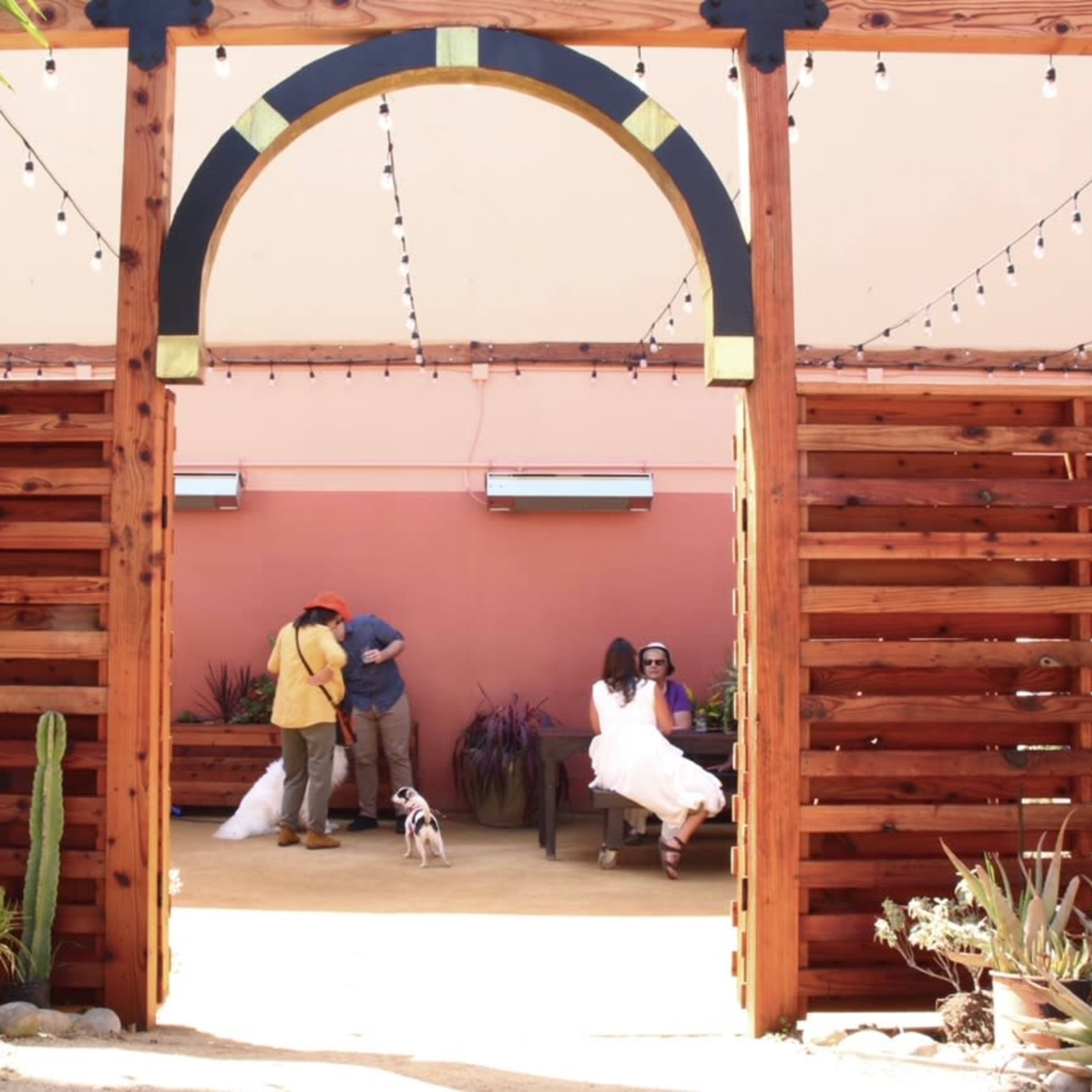A wooden archway frames a patio scene with people mingling, surrounded by potted plants and hanging lights.