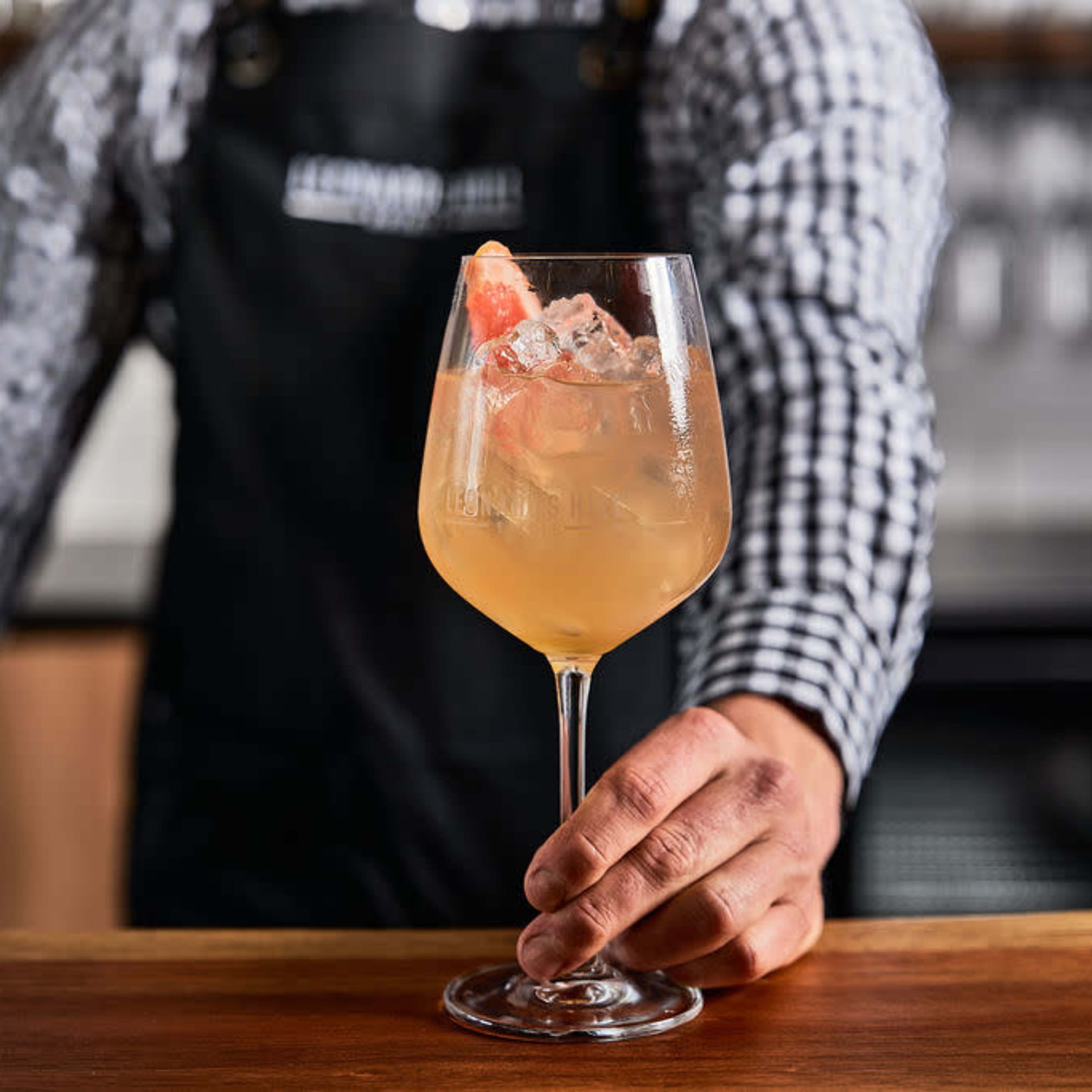 A bartender in a checked shirt and black apron holds a cocktail glass filled with a light-colored drink garnished with ice and a slice of grapefruit.