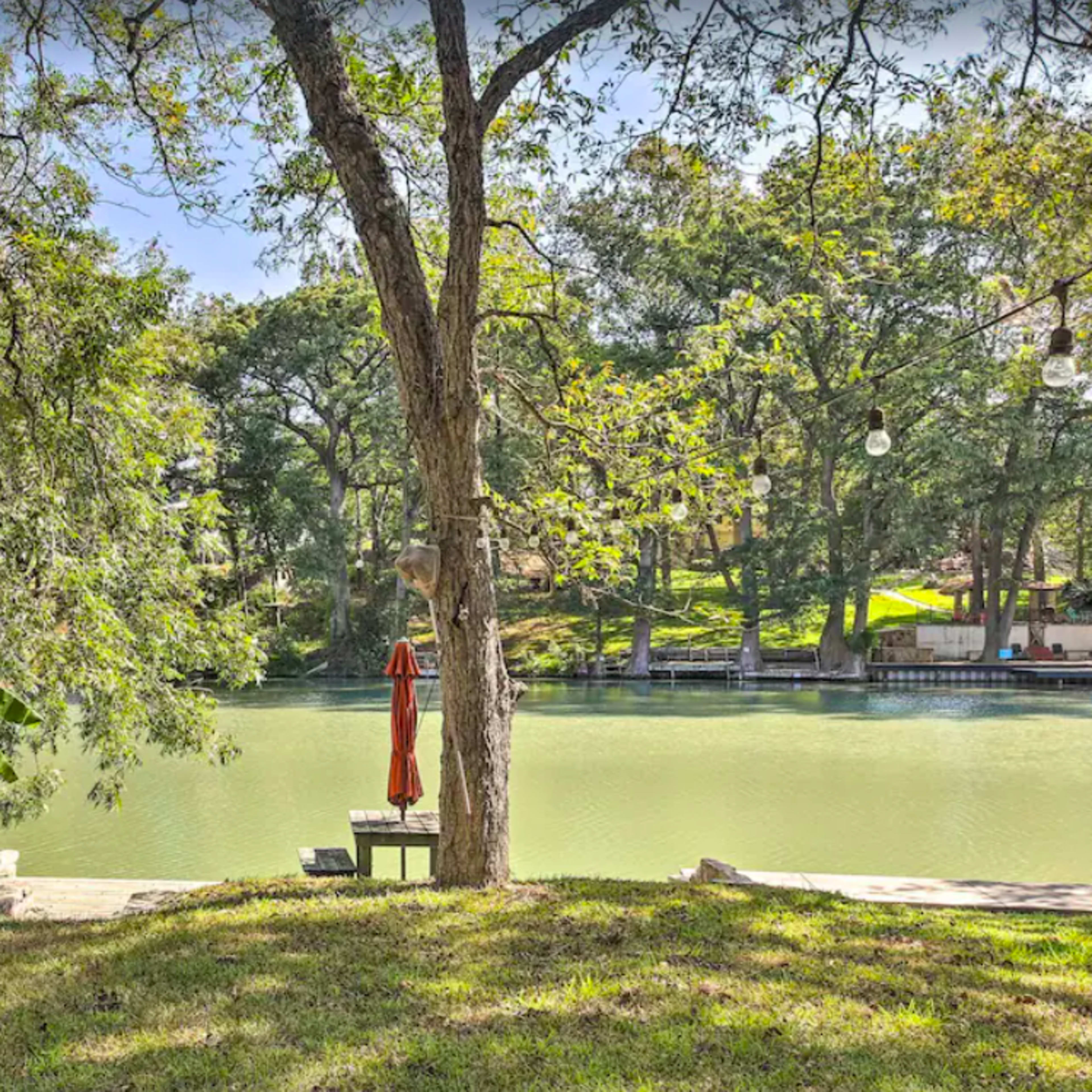 The scene shows a calm river bank framed by trees, with a lone chair and an umbrella positioned near the water's edge.
