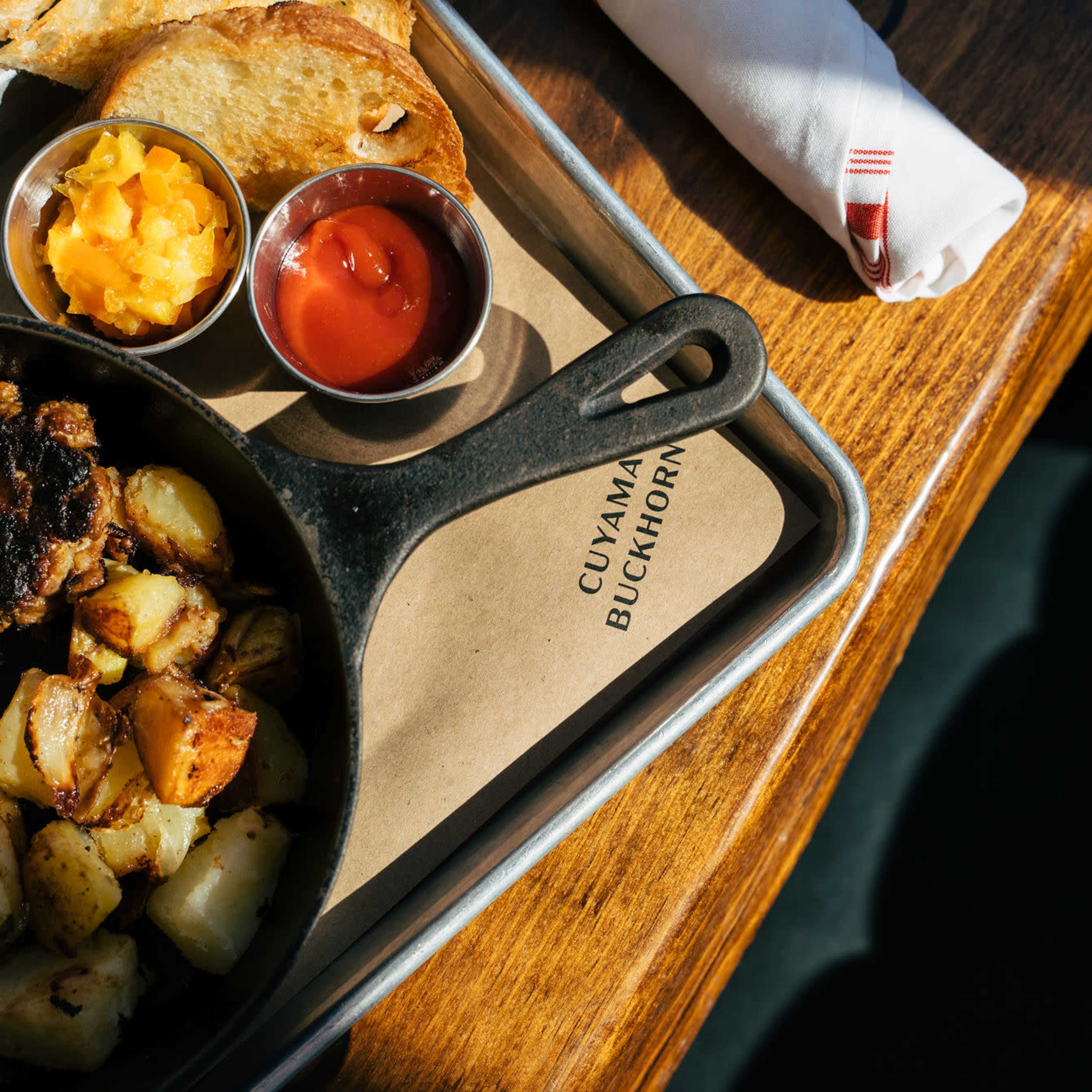 A cast-iron skillet filled with scrambled eggs, grilled sausage, roasted potatoes, and accompanied by cups of chutney and ketchup sits on a wooden table.