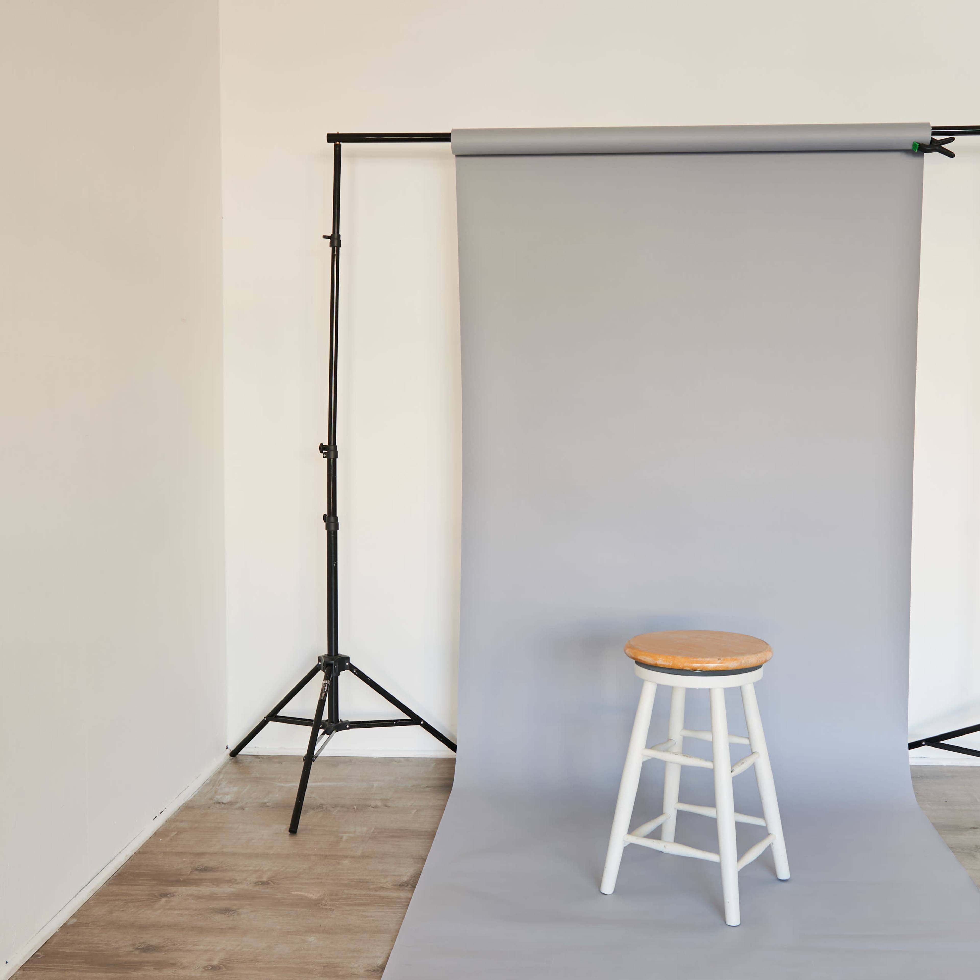 A wooden stool sits in front of a gray backdrop supported by a black frame in a simple studio setting.