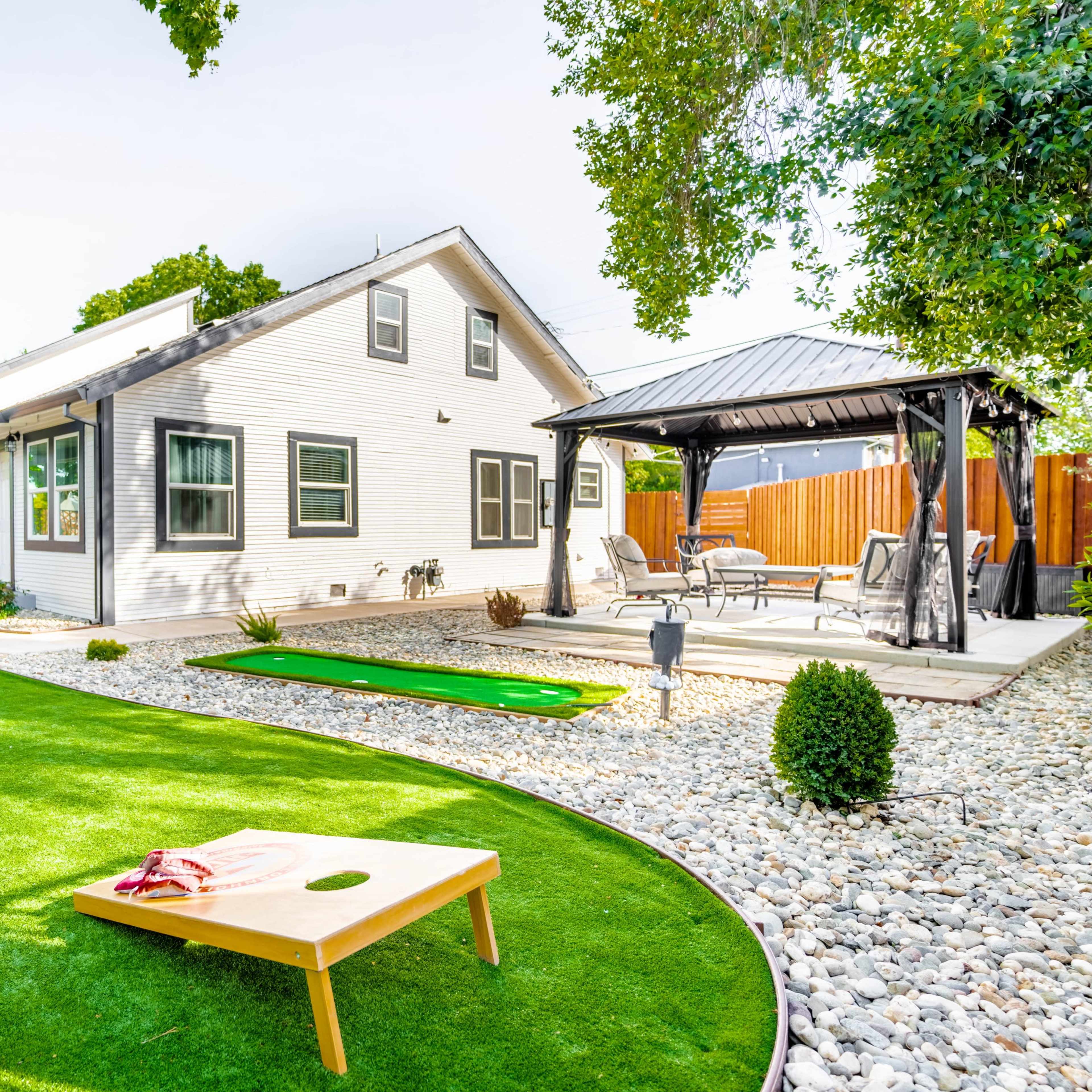 A landscaped backyard featuring a green artificial turf area, a cornhole game set up, a putting green, and a patio with seating.