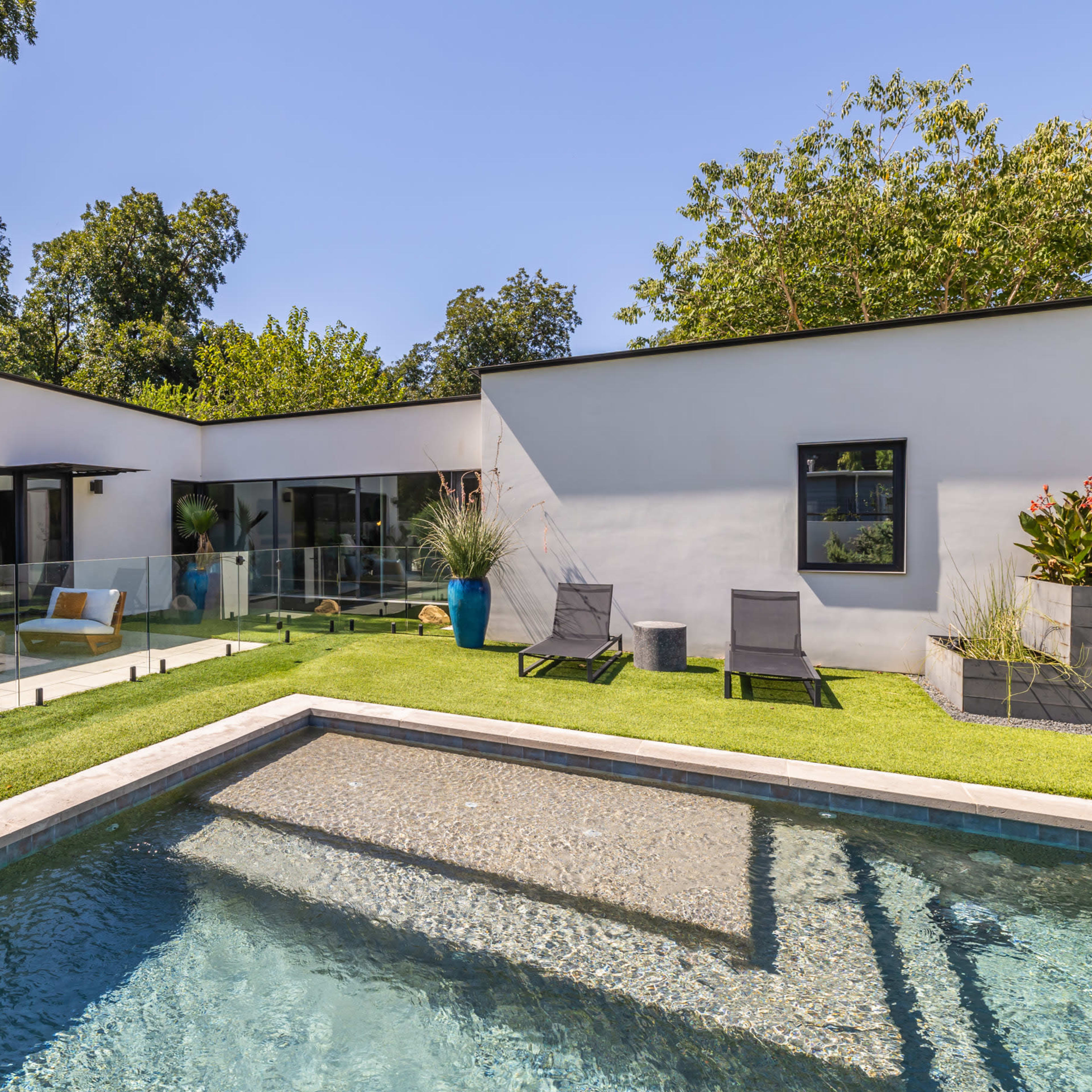 A modern house with large windows surrounding a rectangular swimming pool and a landscaped yard featuring lounge chairs and decorative plants.