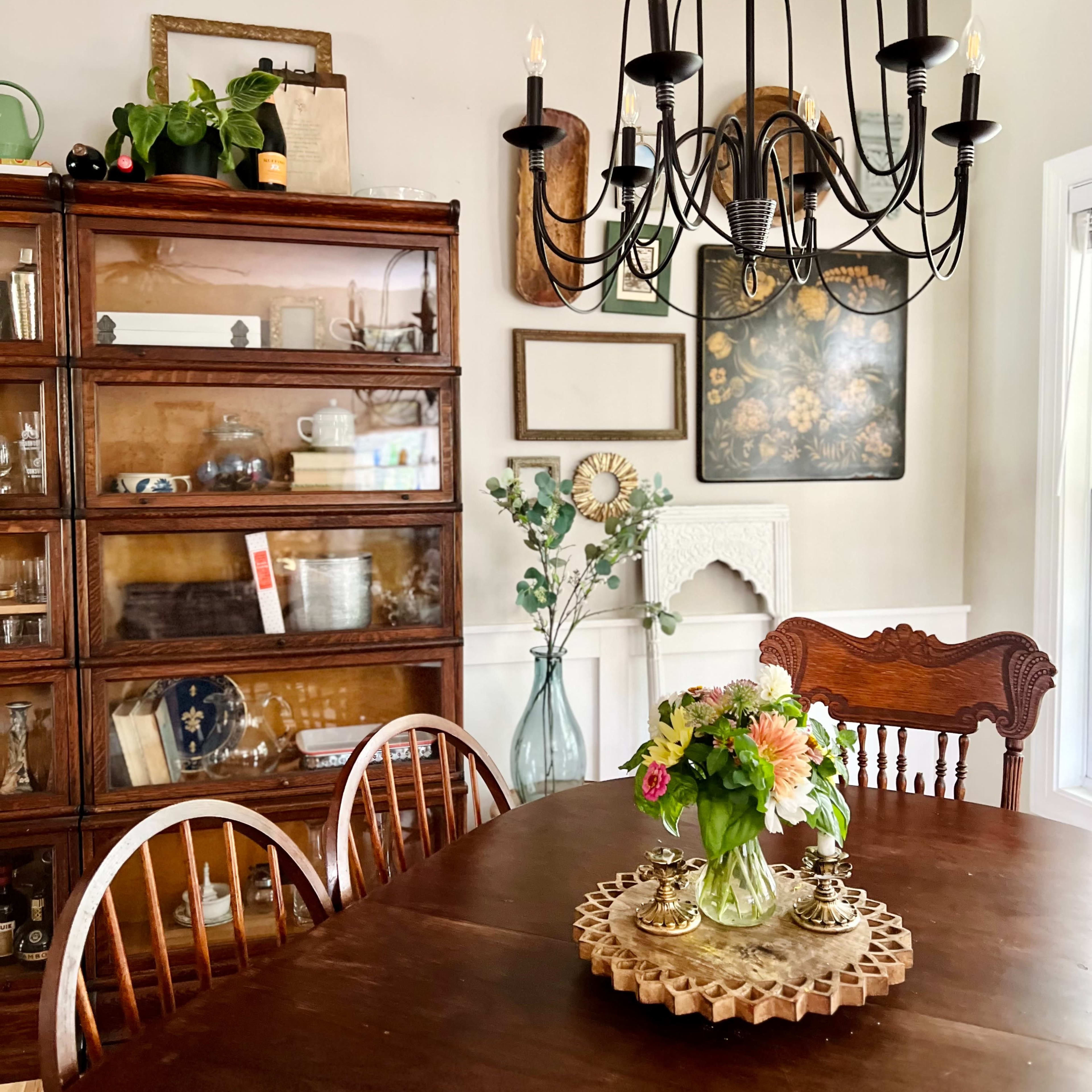 A wooden dining table is set in a well-lit room featuring a chandelier and a cabinet showcasing various items, along with a floral centerpiece.